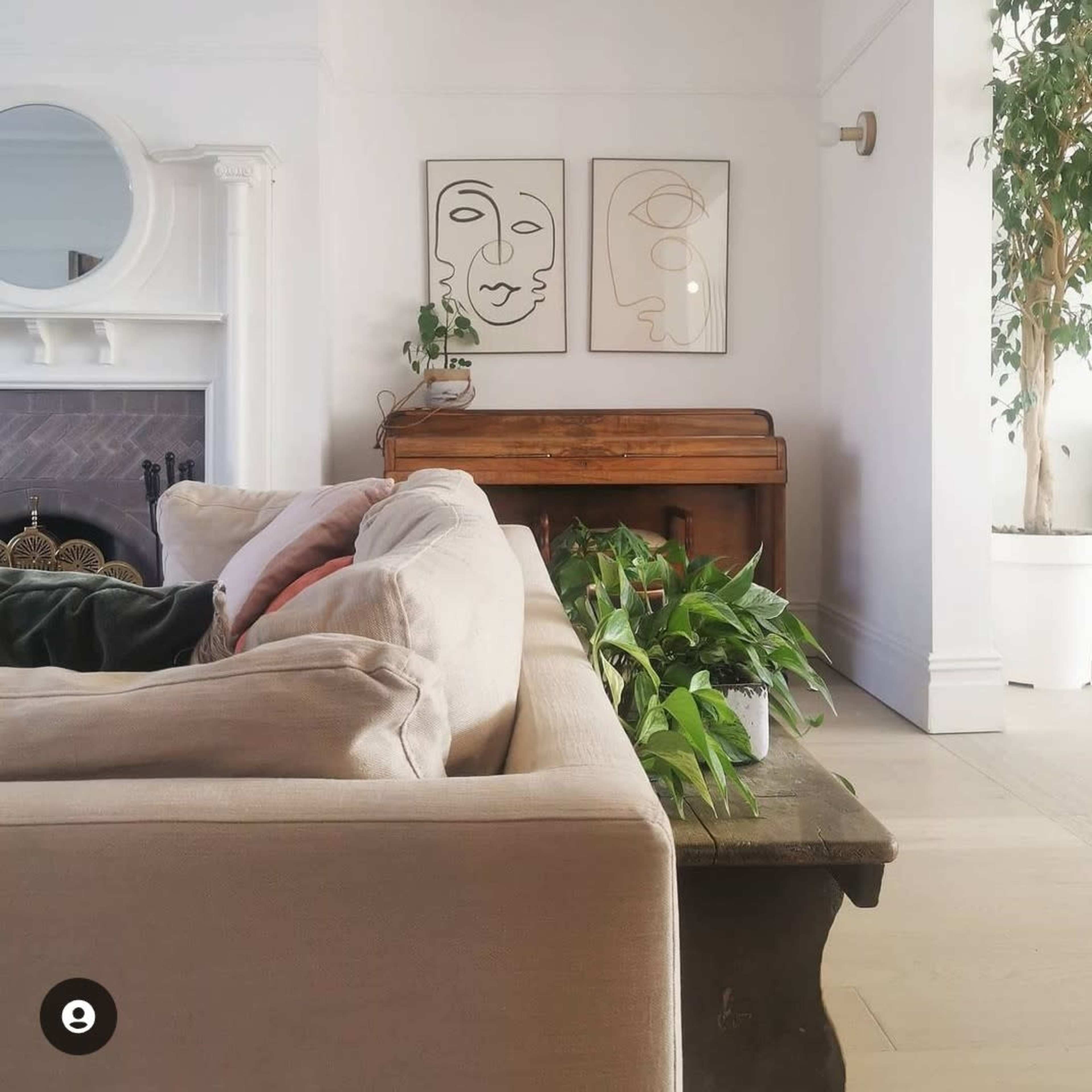 A living room with a light-colored sofa, a wooden piano, two framed line art portraits on the wall, and green plants along a table.