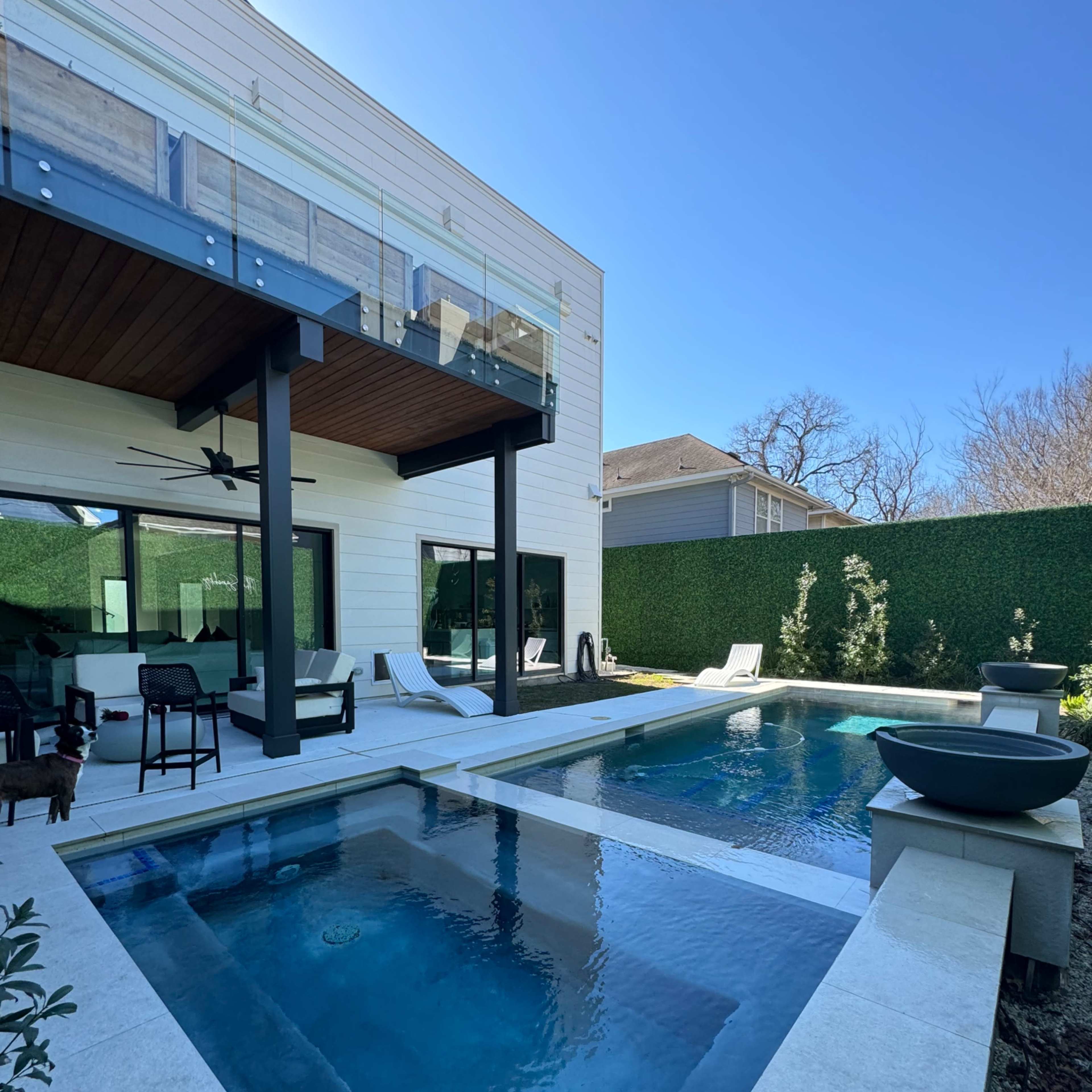 A modern outdoor area featuring a pool, a patio with seating, and lush green hedges surrounding the space.
