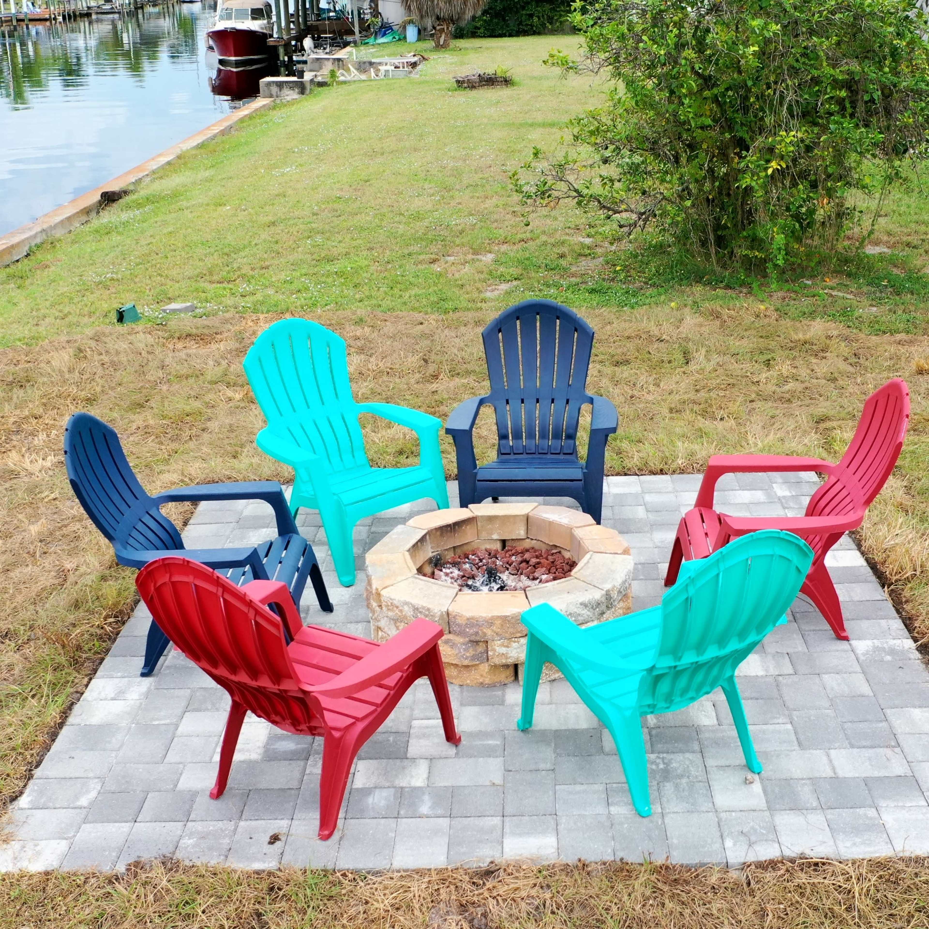 A fire pit surrounded by six colorful Adirondack chairs is set on a grassy area beside a body of water.
