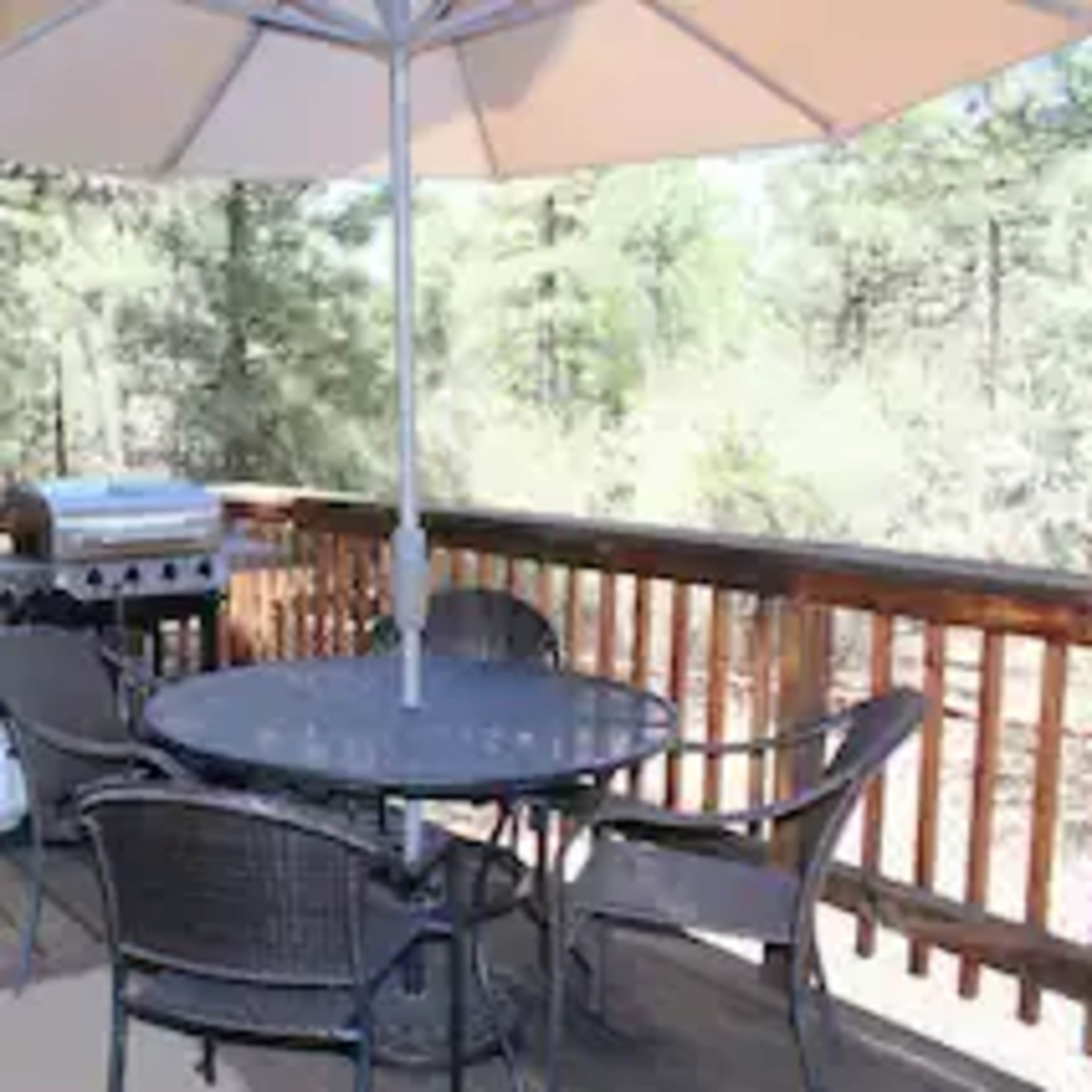 A dining table with four chairs and an umbrella is set on a wooden deck beside a grill and a small shed, surrounded by trees.