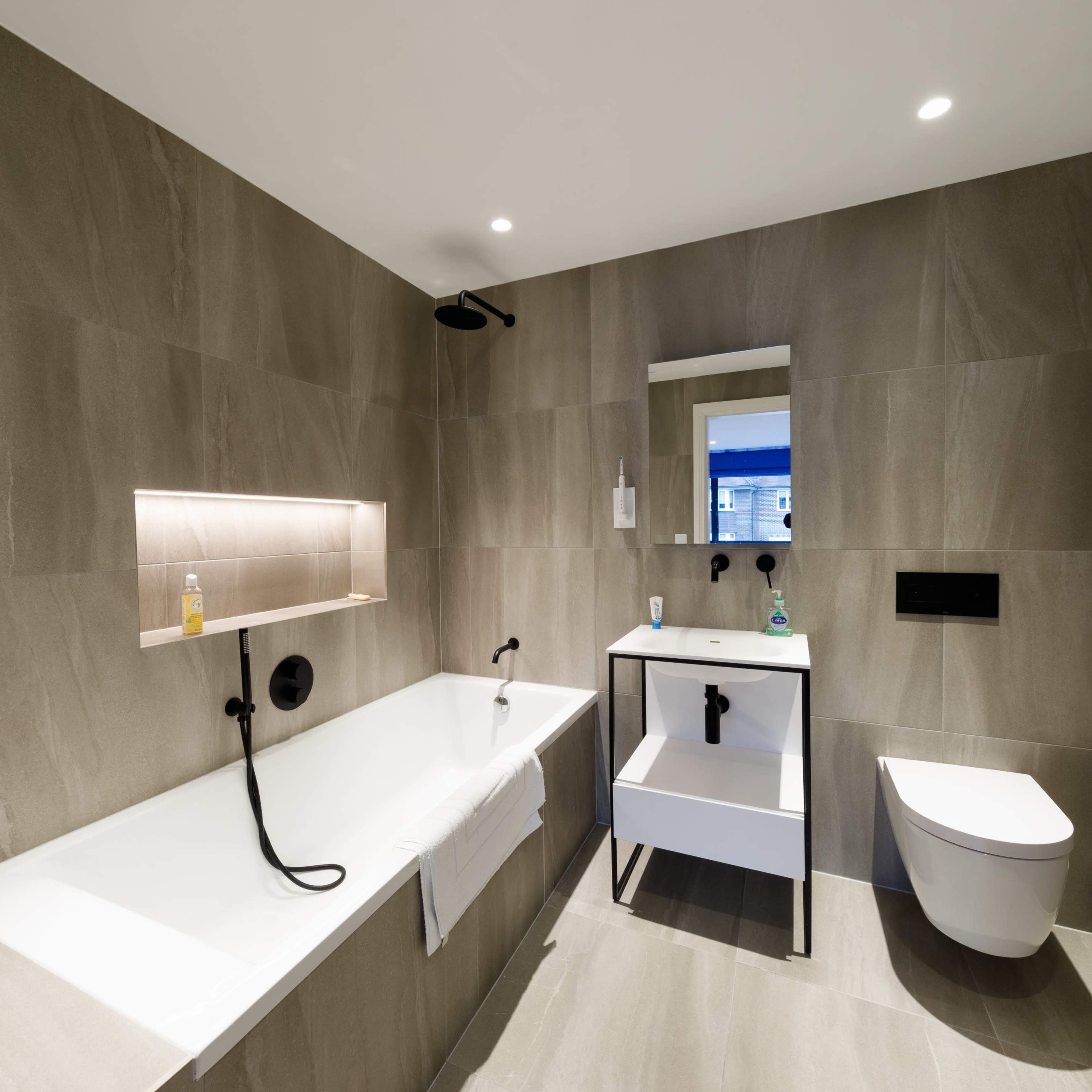 A modern bathroom featuring a bathtub, a wall-mounted sink with storage, and a toilet, all surrounded by gray tiled walls.