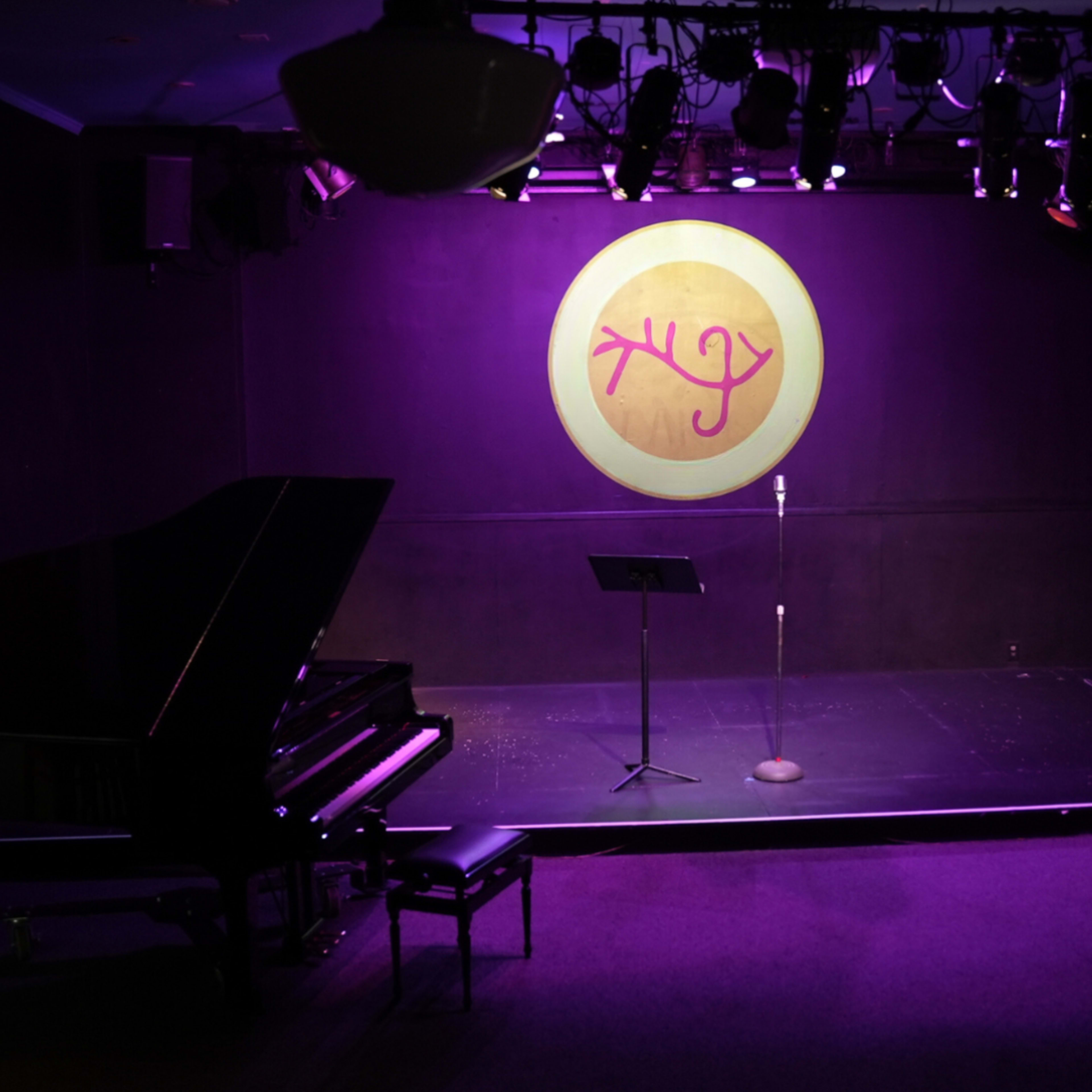 An empty performance stage with a grand piano on the left, a microphone stand centered, and a circular logo illuminated on the back wall.