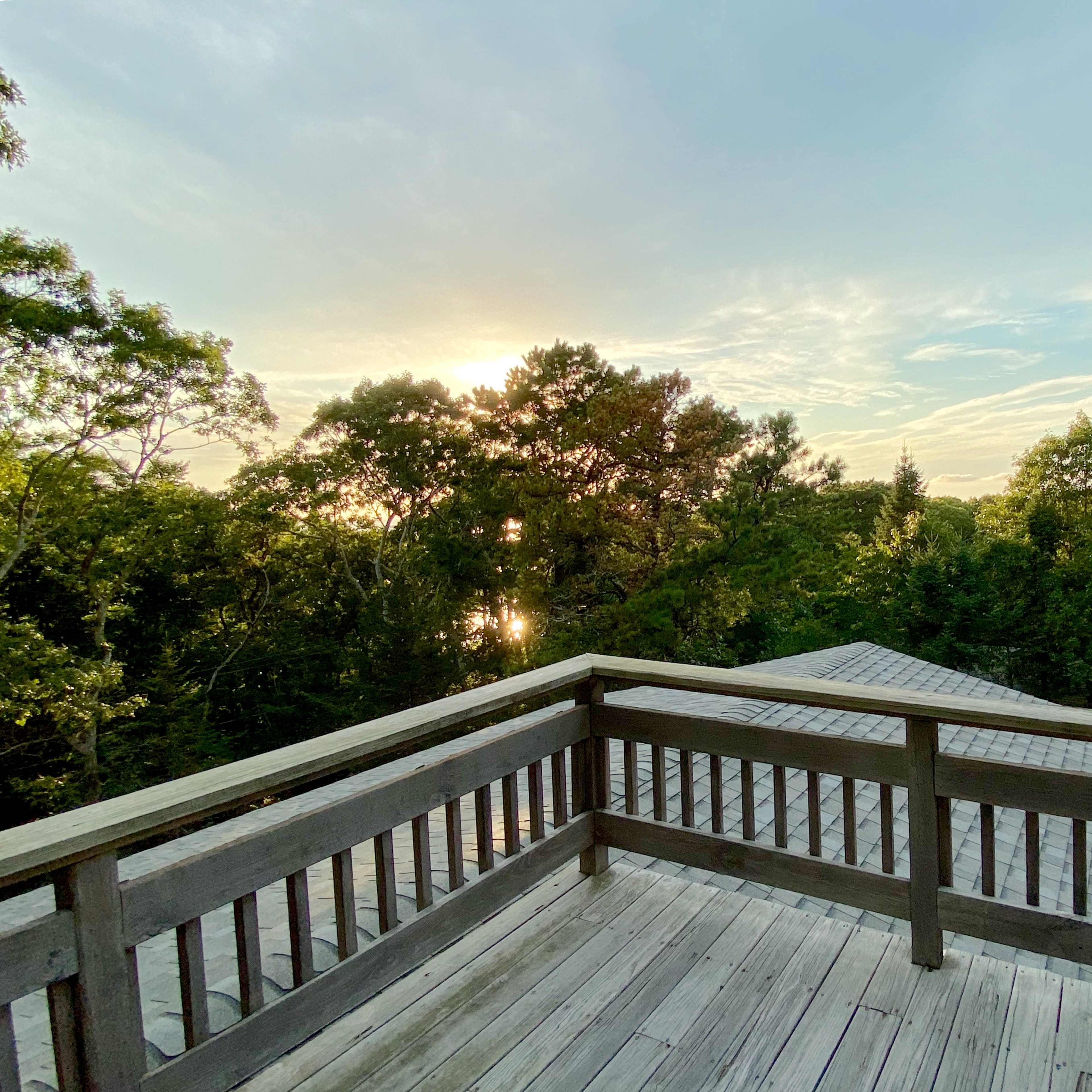 A wooden deck overlooking a wooded area with a view of a sunset in the background.