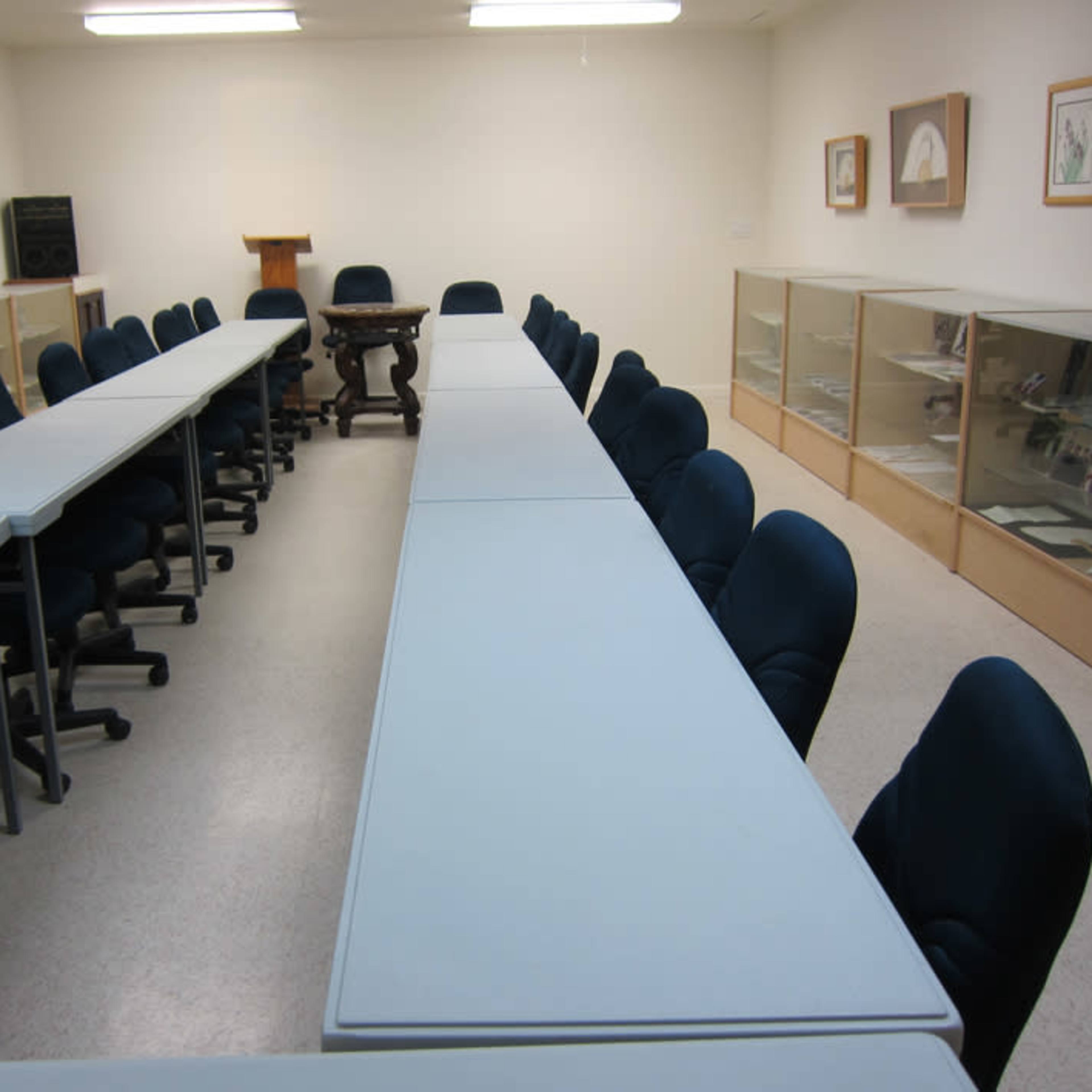 A meeting room features several blue chairs arranged around long, white tables, with display cases and framed pictures along the walls.