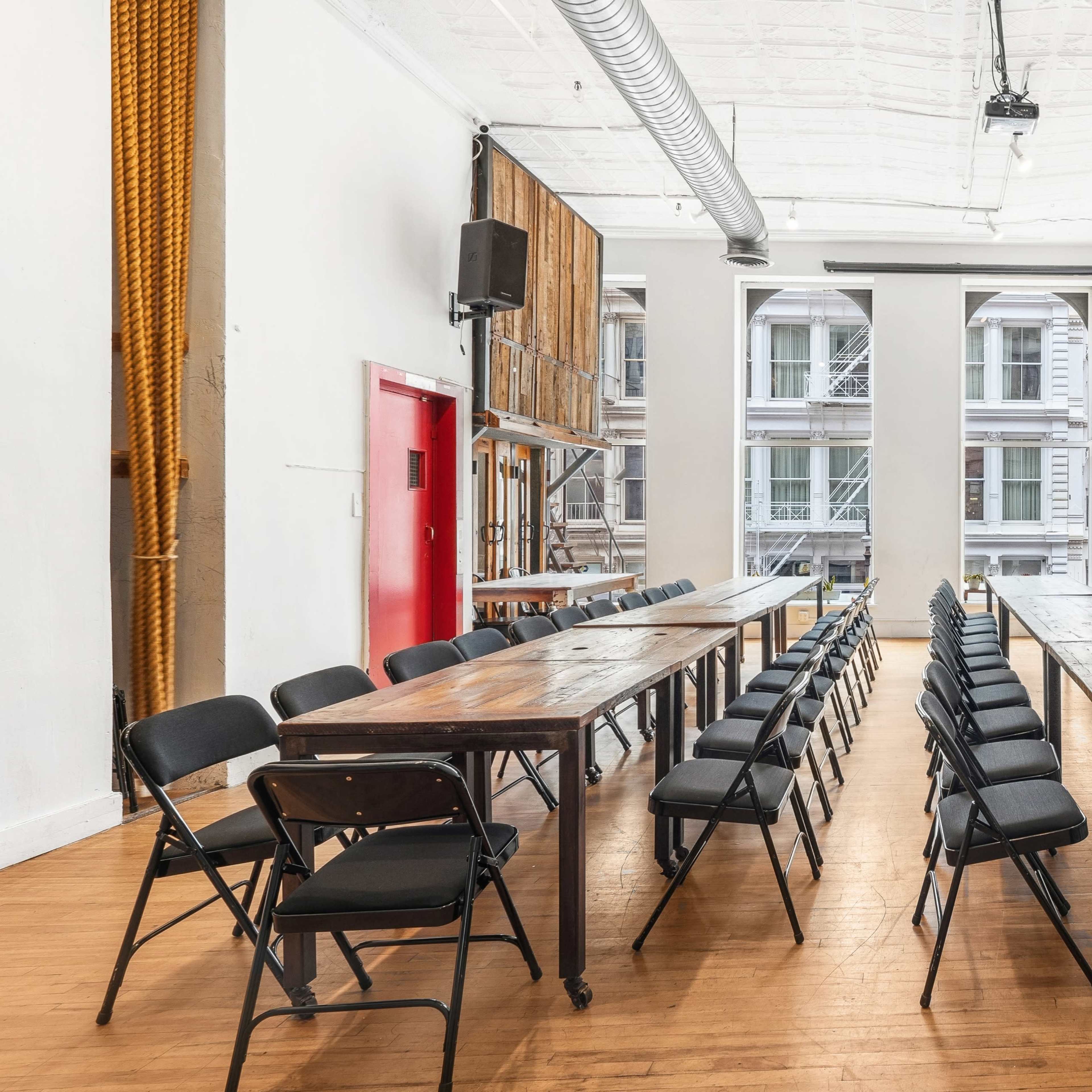 A spacious room features two long wooden tables arranged in parallel with black folding chairs positioned around them, illuminated by natural light from large windows.