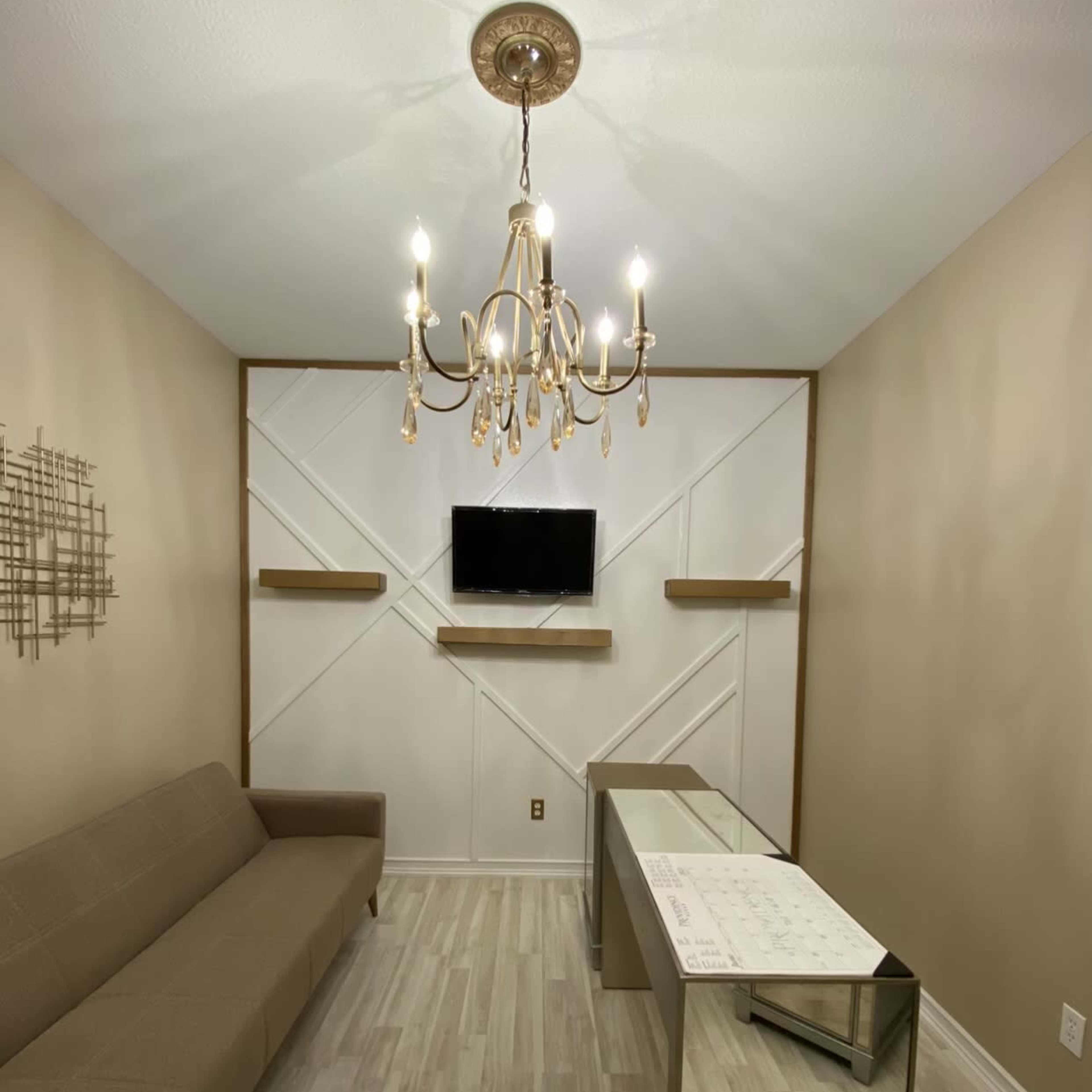 A small, elegantly designed room featuring a beige sofa, a glass desk, a chandelier, and a television mounted on the wall.