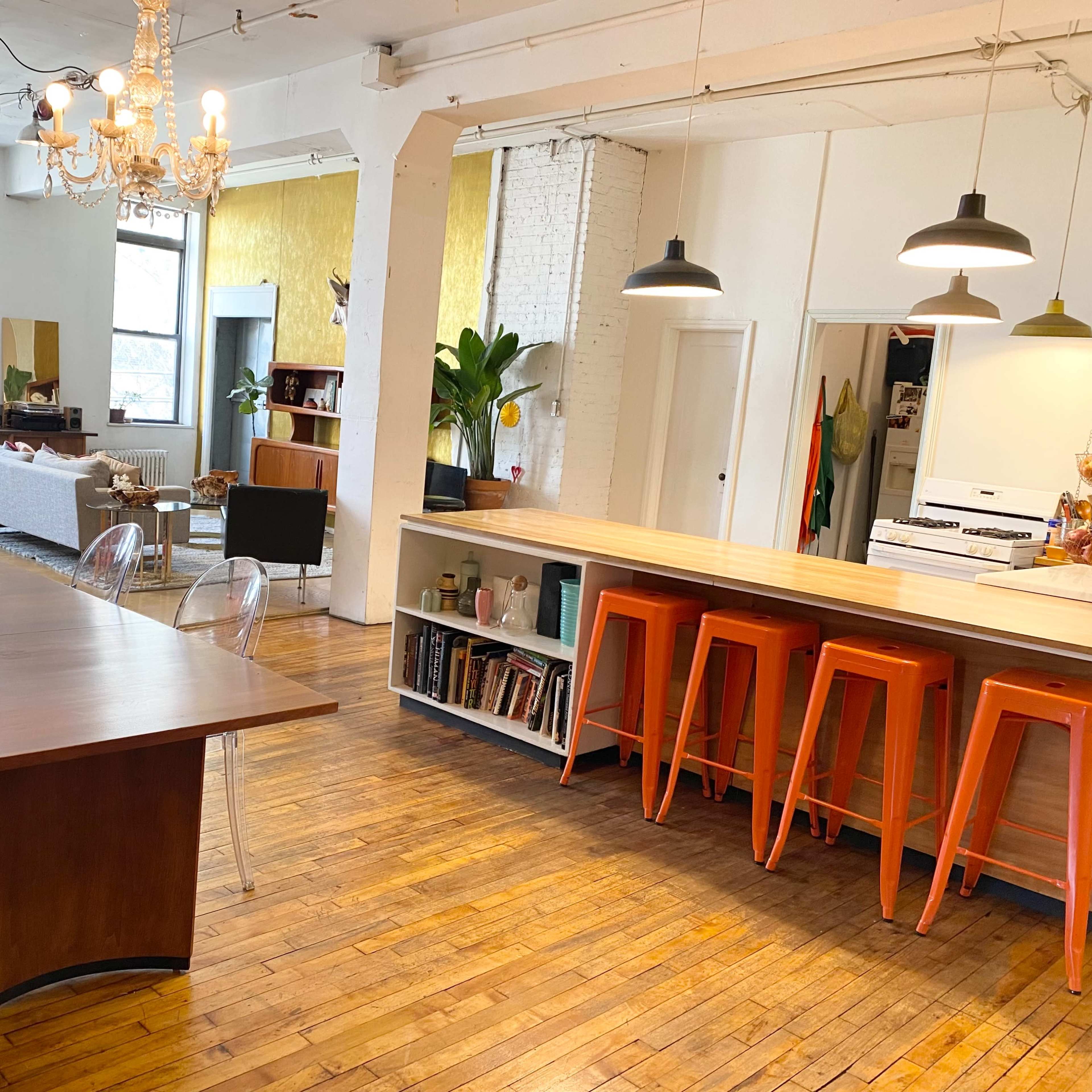 A bright, open living space featuring a wooden dining table, a kitchen with orange stools, and large windows allowing natural light to fill the room.