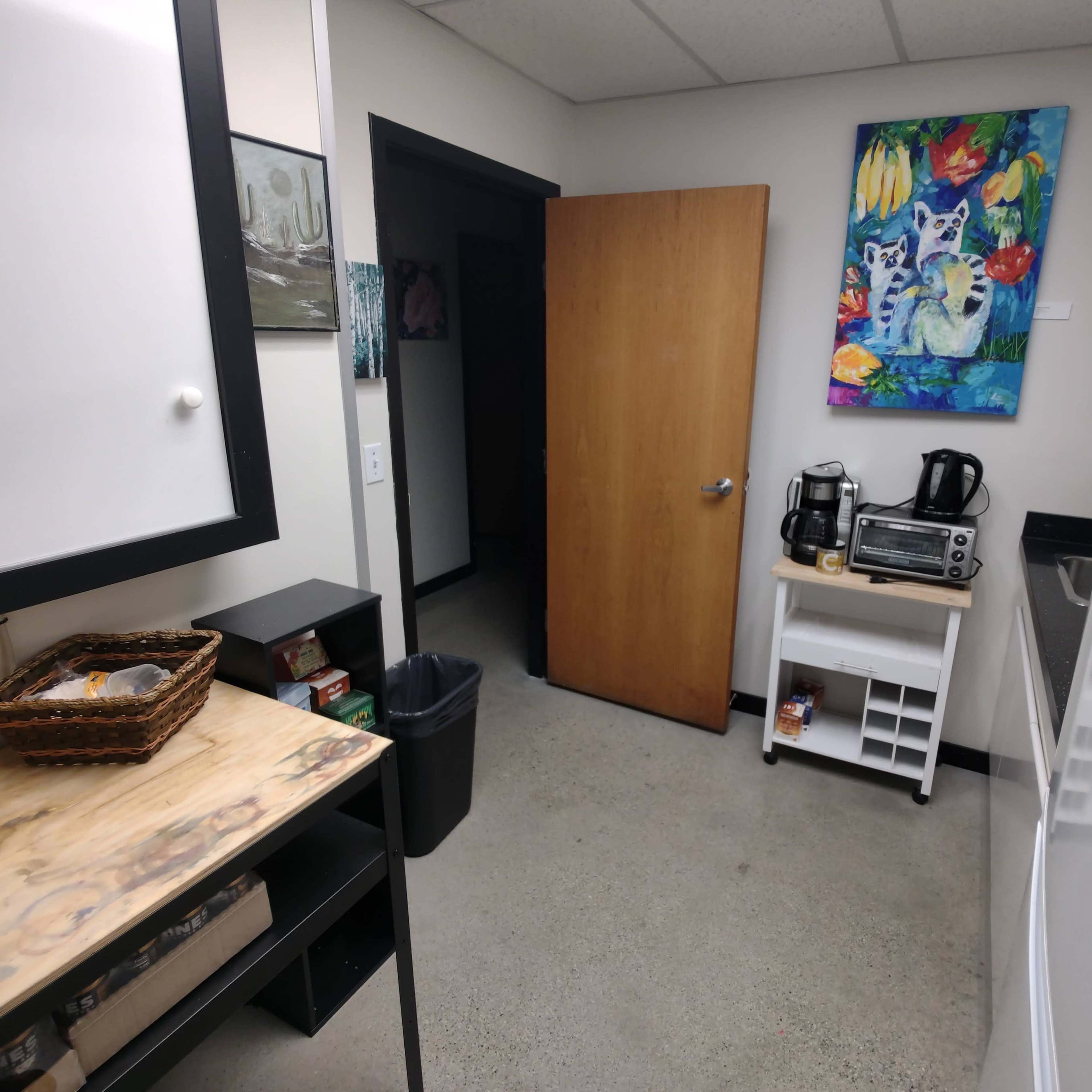 A break room with a wooden shelf, a trash can, a refrigerator, a coffee machine, and a colorful painting on the wall.