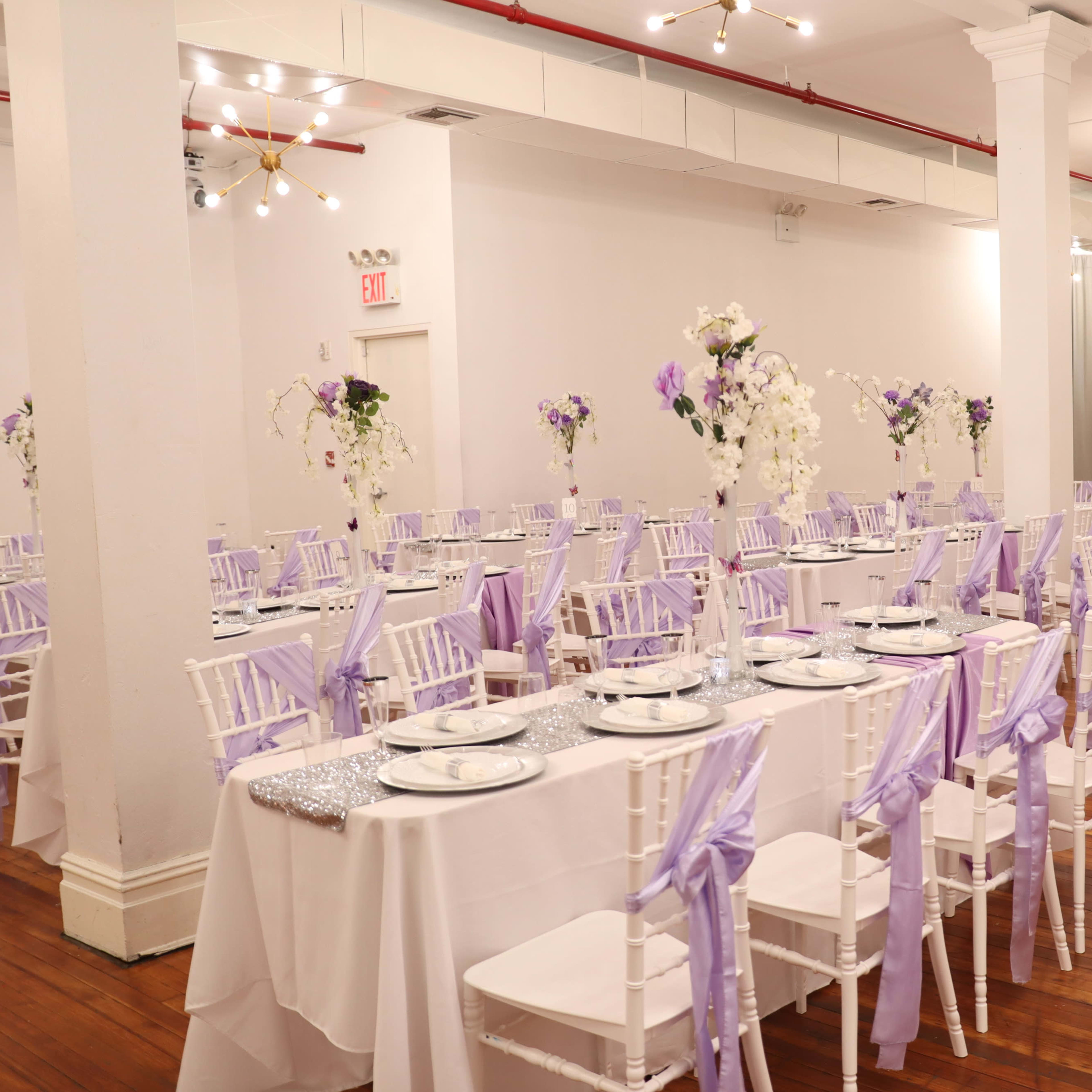 An elegantly arranged banquet hall with tables set for an event, featuring white tablecloths, lavender accents, and floral centerpieces.