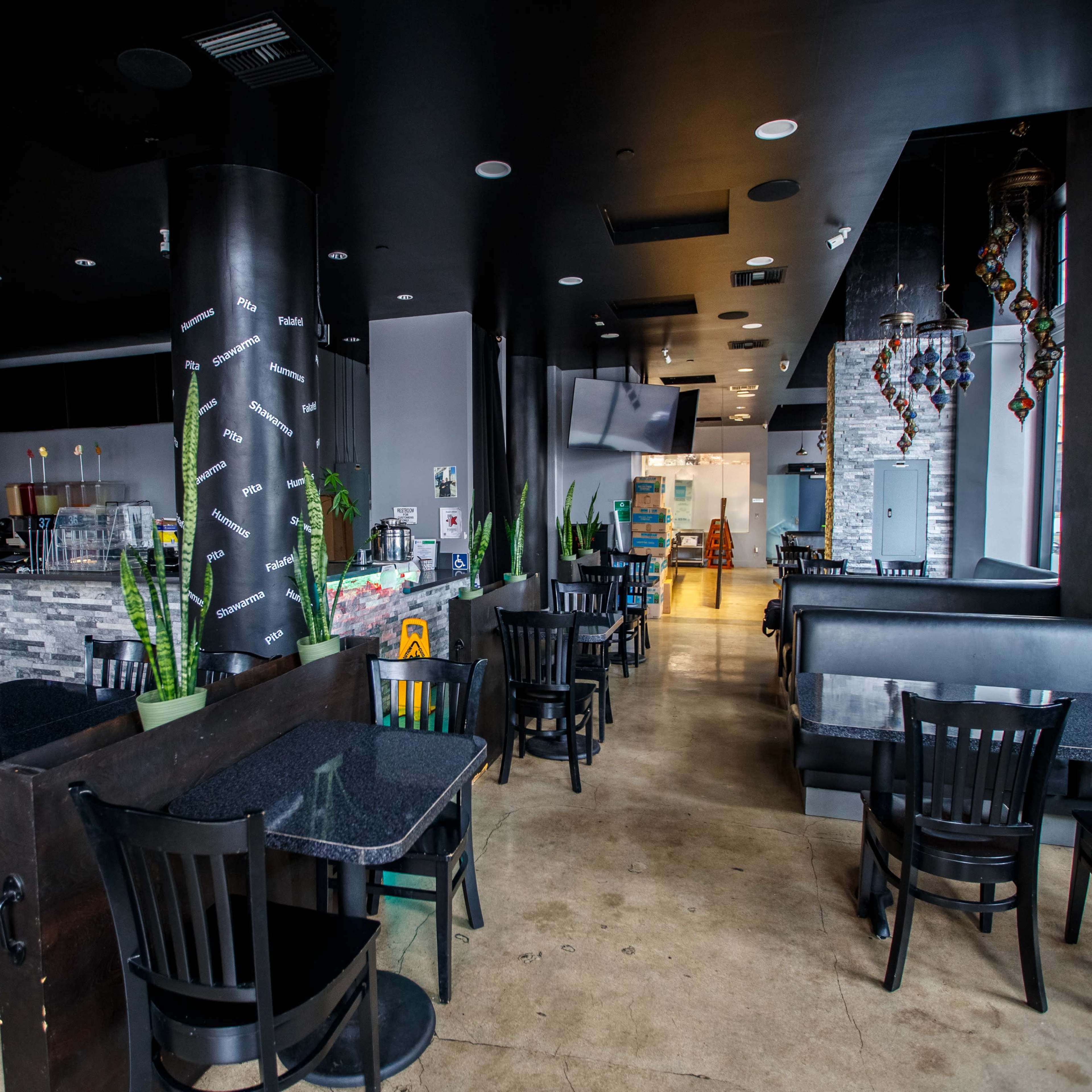 A modern restaurant interior with black tables and chairs, potted plants, and a counter area at the back.