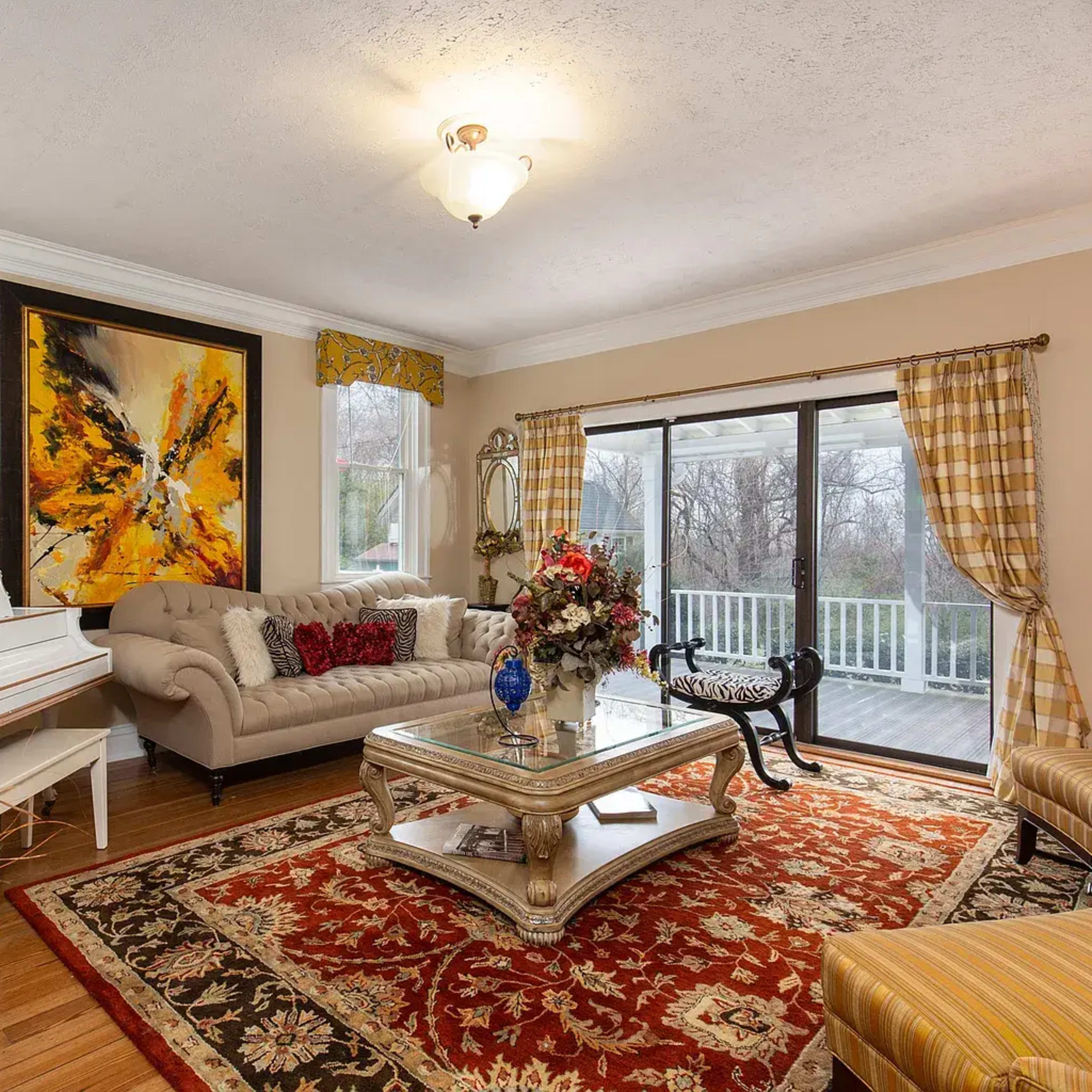 The room features a beige color scheme with a piano, a decorative coffee table, a large area rug, and large windows overlooking a porch.