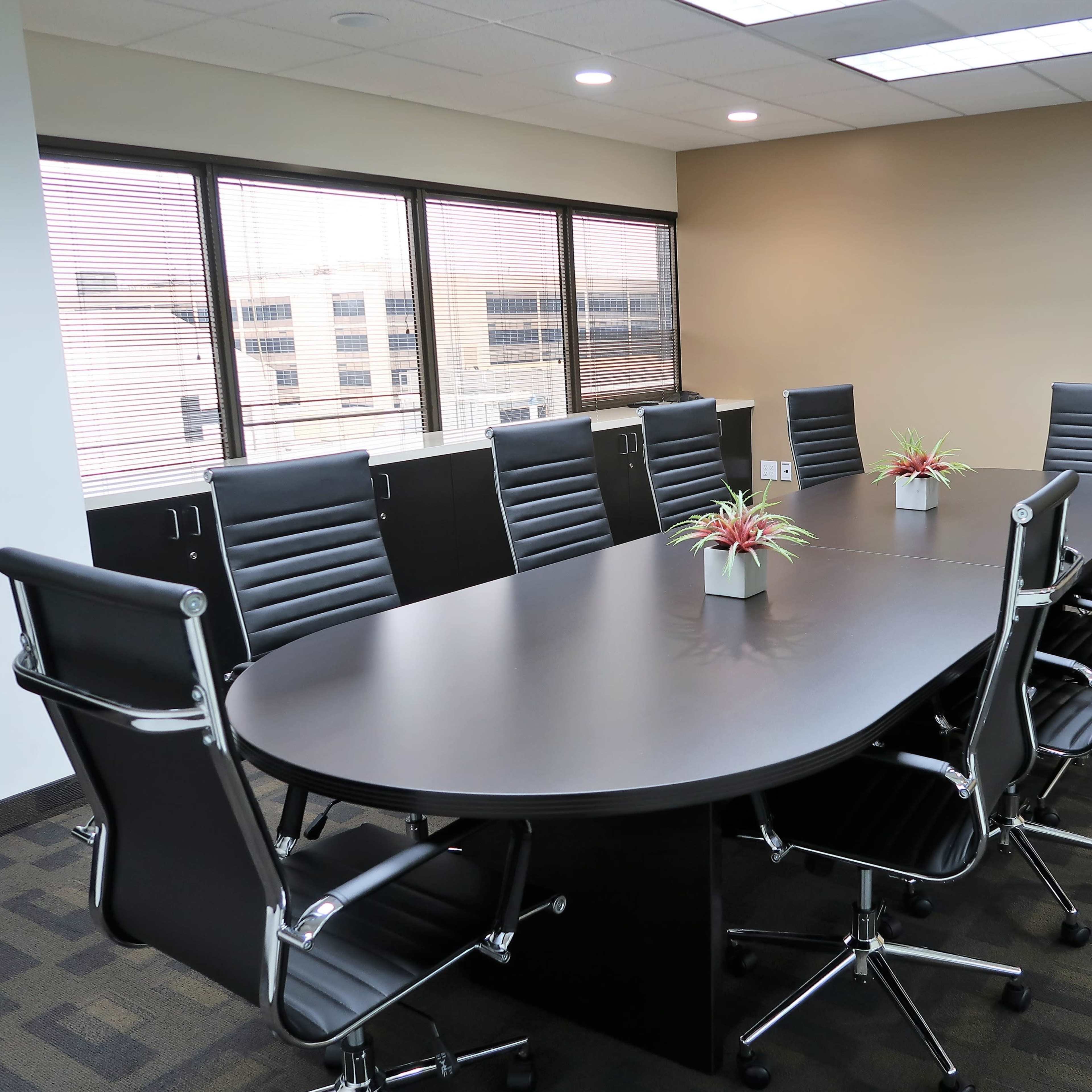 A modern conference room features a large oval table surrounded by ergonomic chairs and plants, with large windows providing a view of the city.
