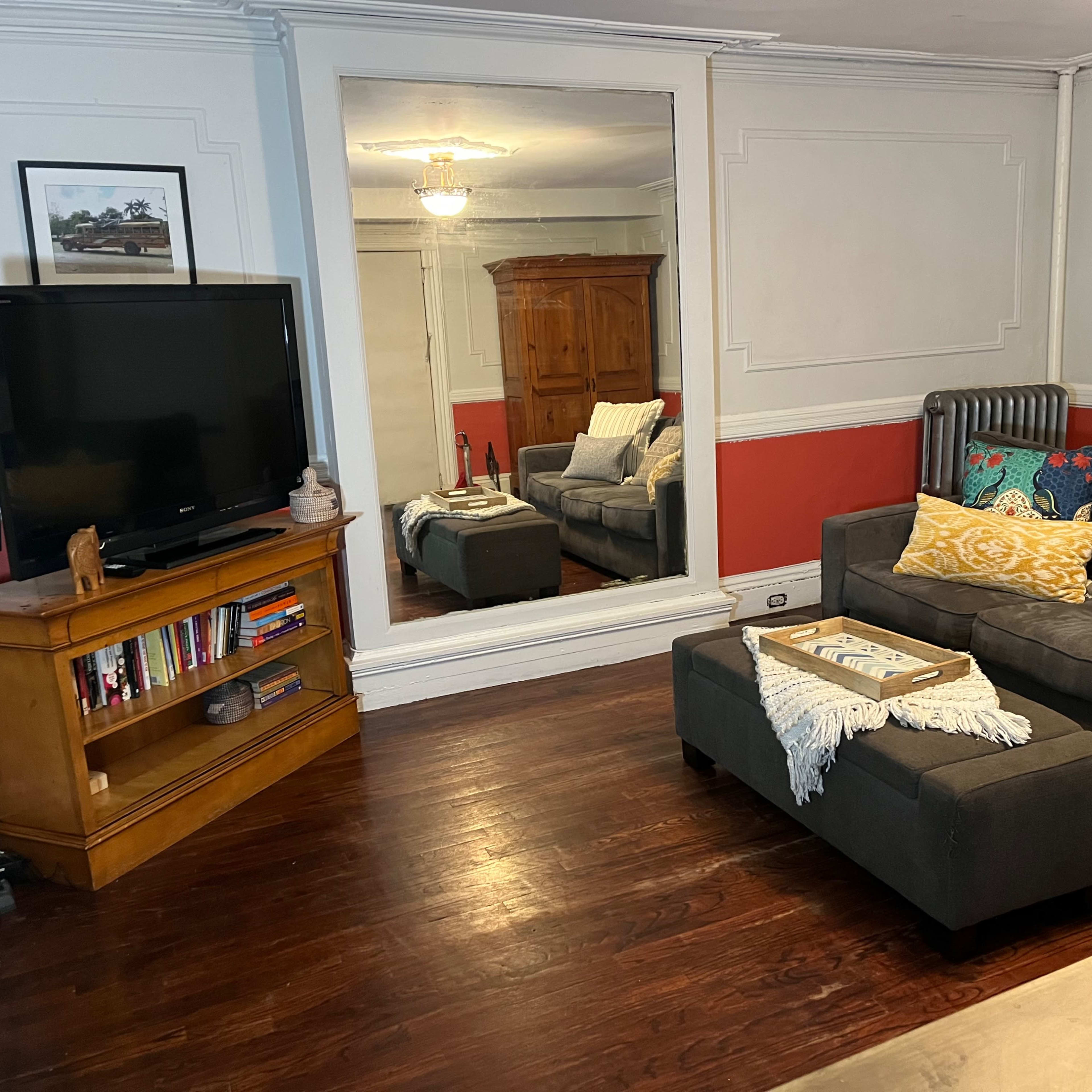 The living room features a gray sectional sofa with decorative pillows, a wooden TV stand, a large mirror, and hardwood flooring.