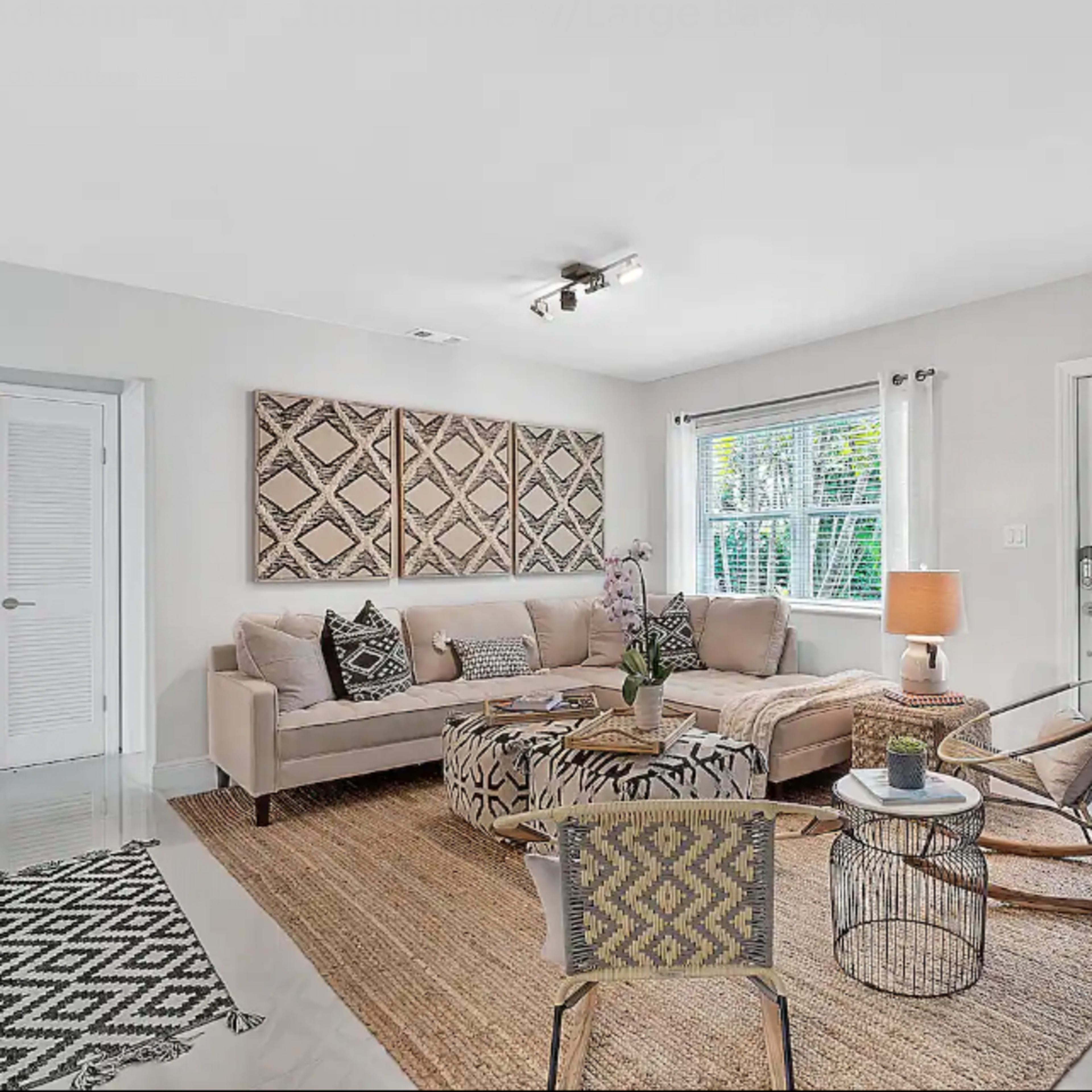 A modern living room with a beige sofa, patterned wall art, a round coffee table, and a large window, complemented by a potted plant and a woven rug.