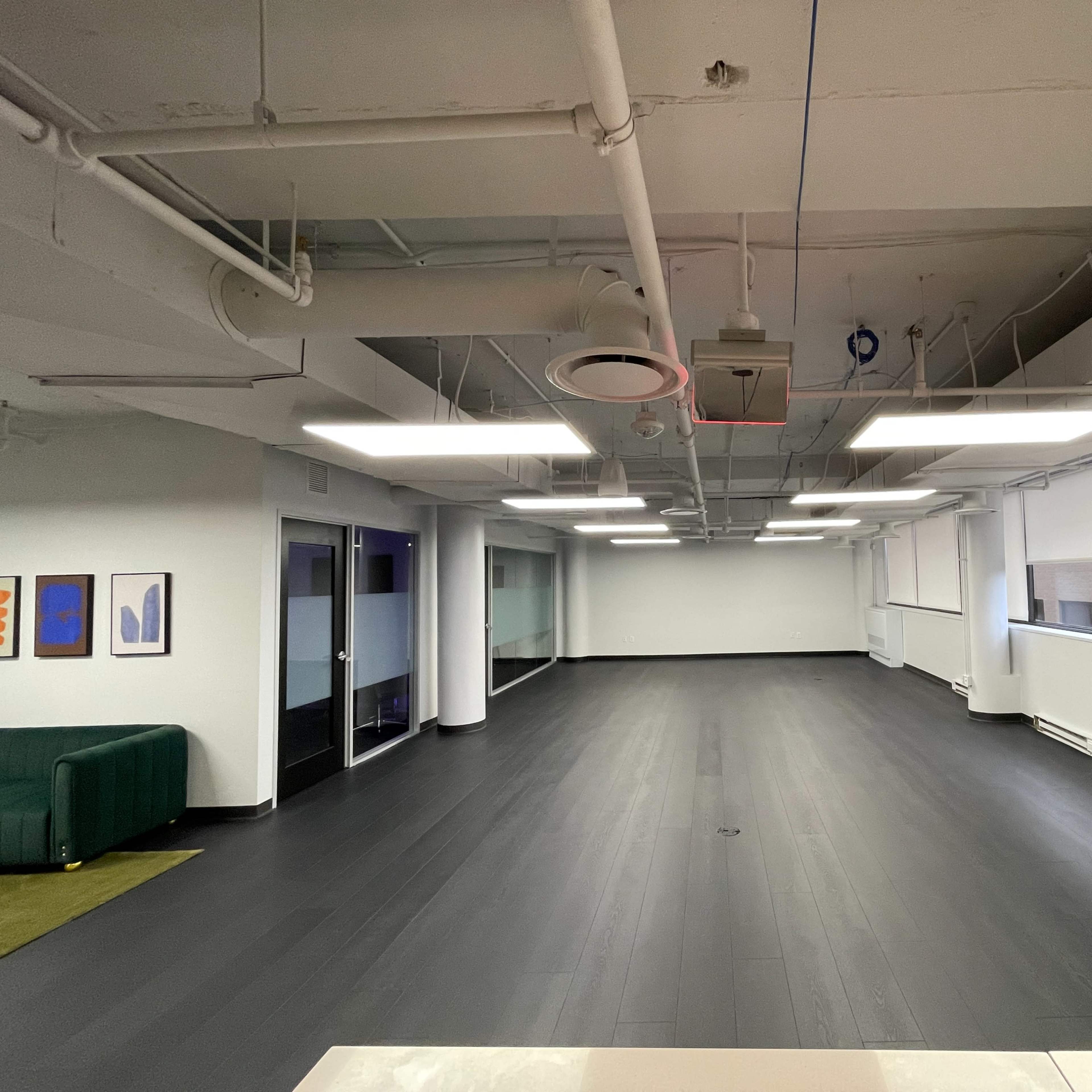 A spacious, empty office with dark wooden floors and overhead lighting, featuring large windows along the walls.