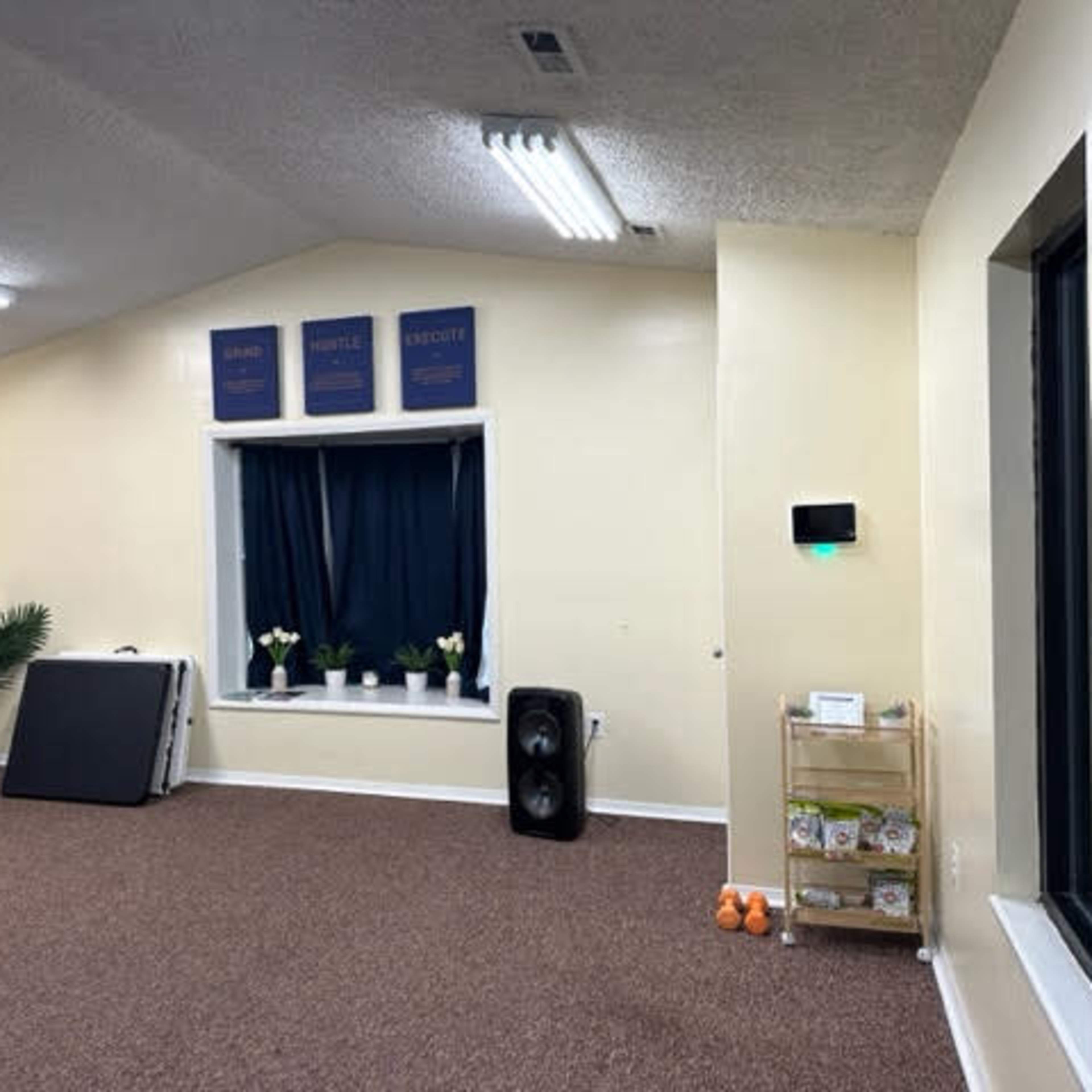 The interior of a room features a carpeted floor, a window with plants, a speaker, and various furniture, including a couch and a rolling cart.