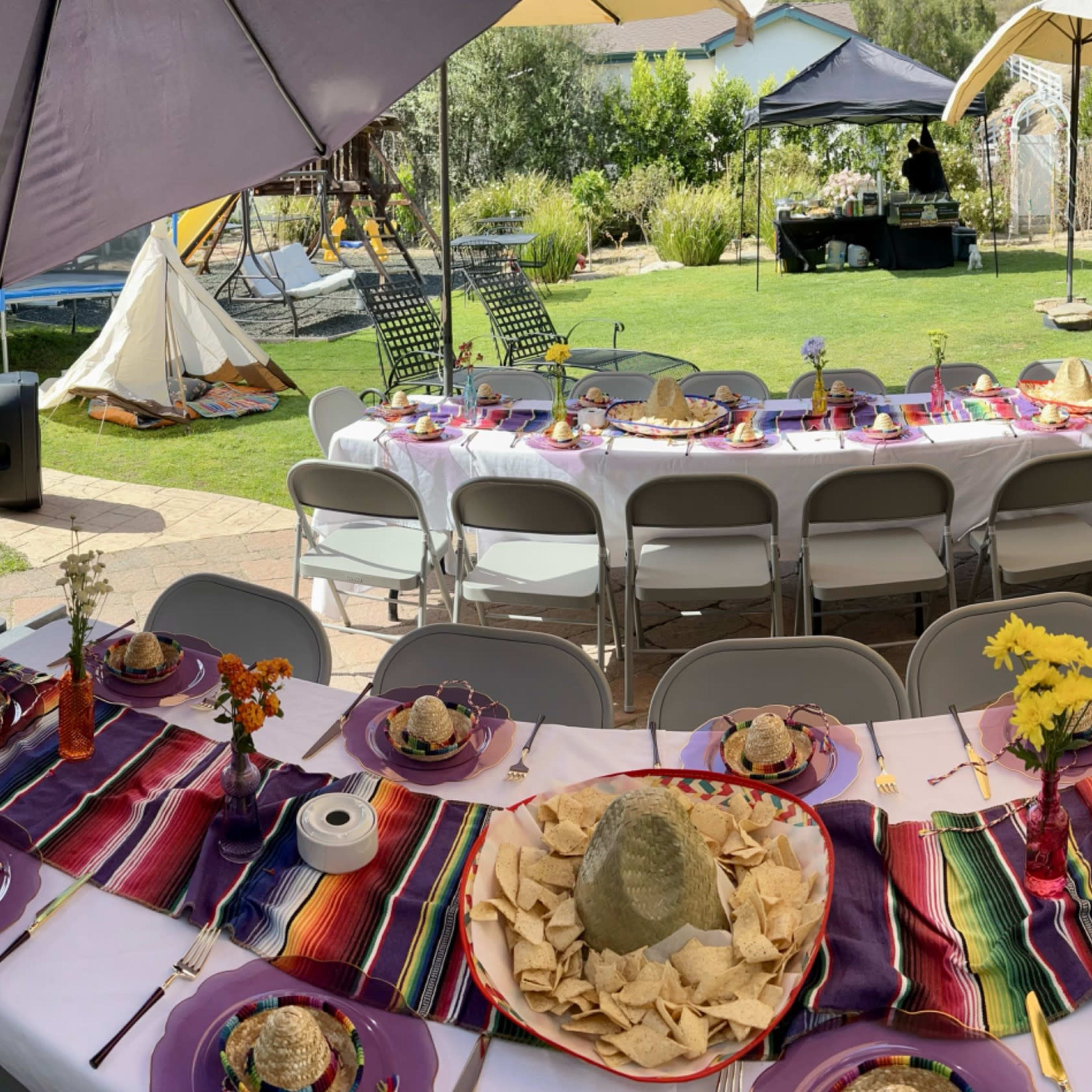 A long table is set for an outdoor gathering, adorned with colorful table runners and plates of food, surrounded by a lush garden.