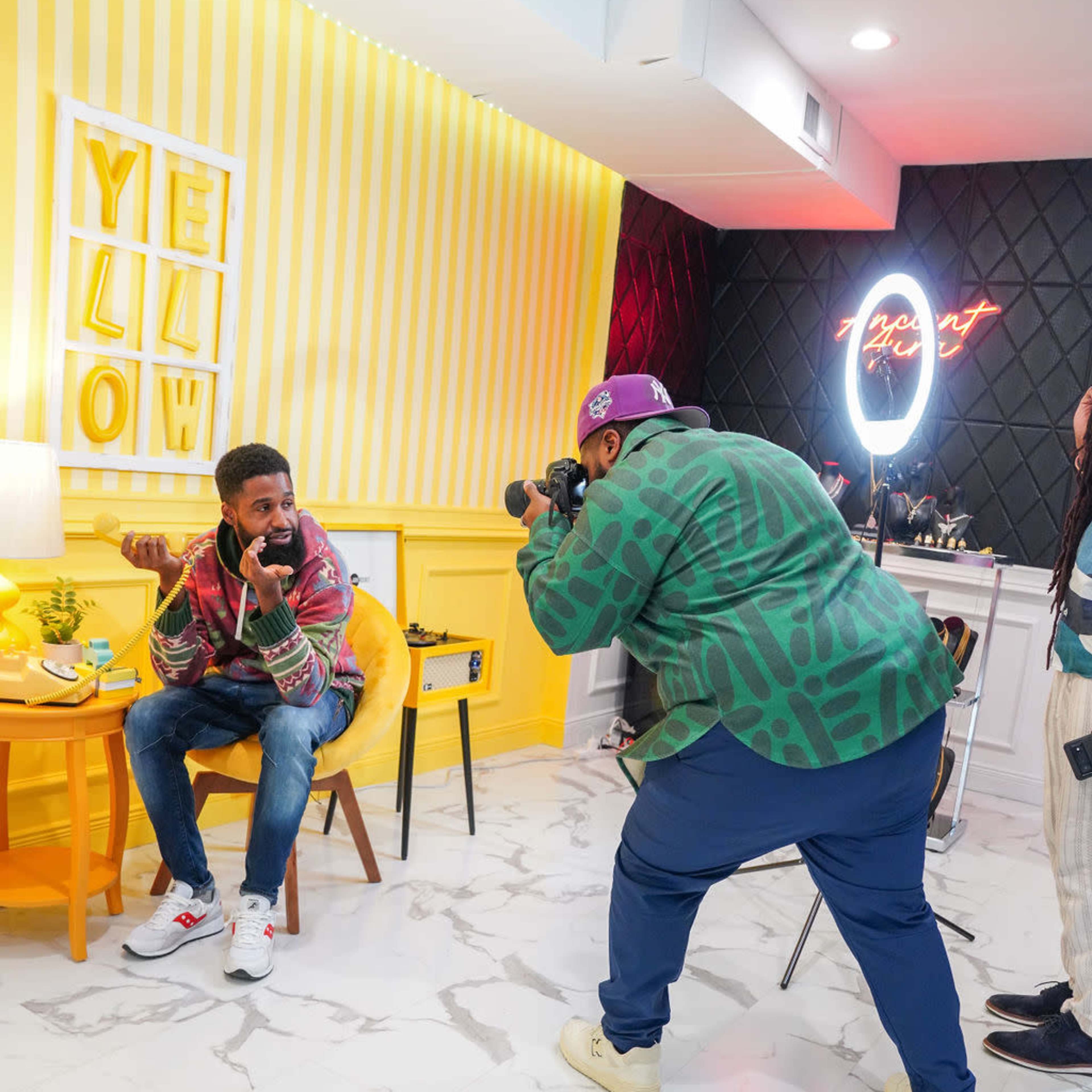 A person is seated on a yellow chair in a brightly decorated room while another person photographs them.