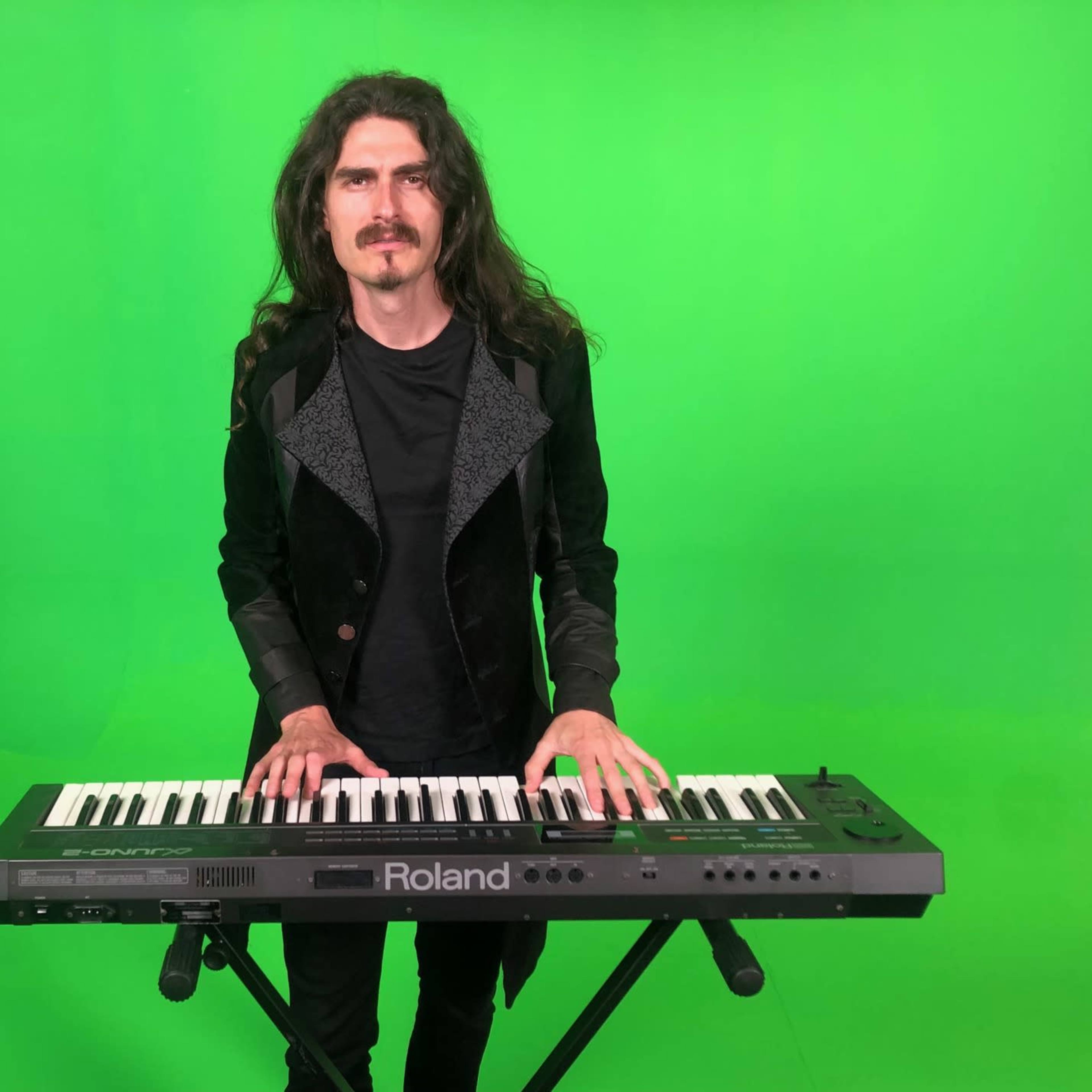 A man with long hair plays a keyboard against a green backdrop.