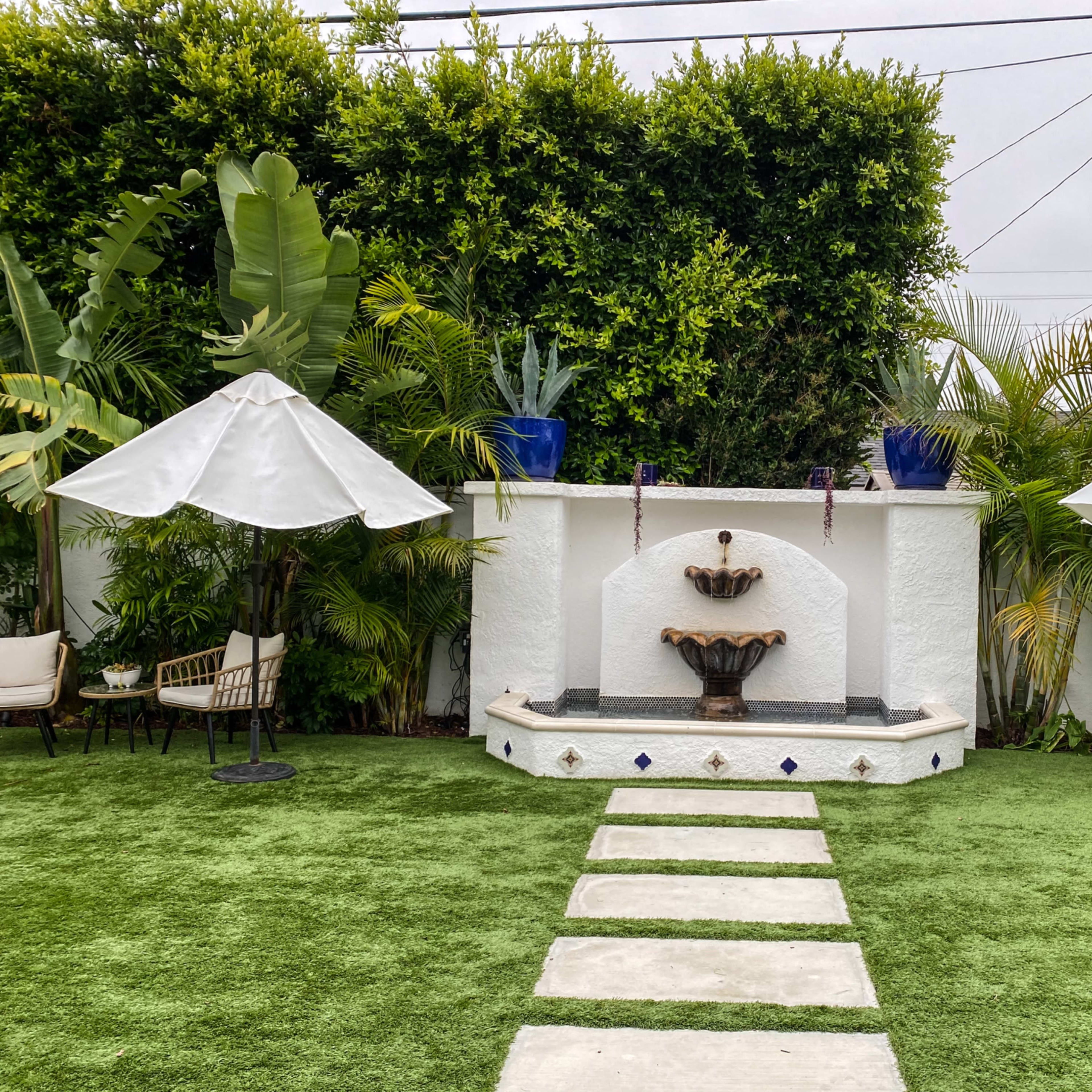 A landscaped garden features a tiered fountain, surrounded by tropical plants and two white umbrellas over seating areas.