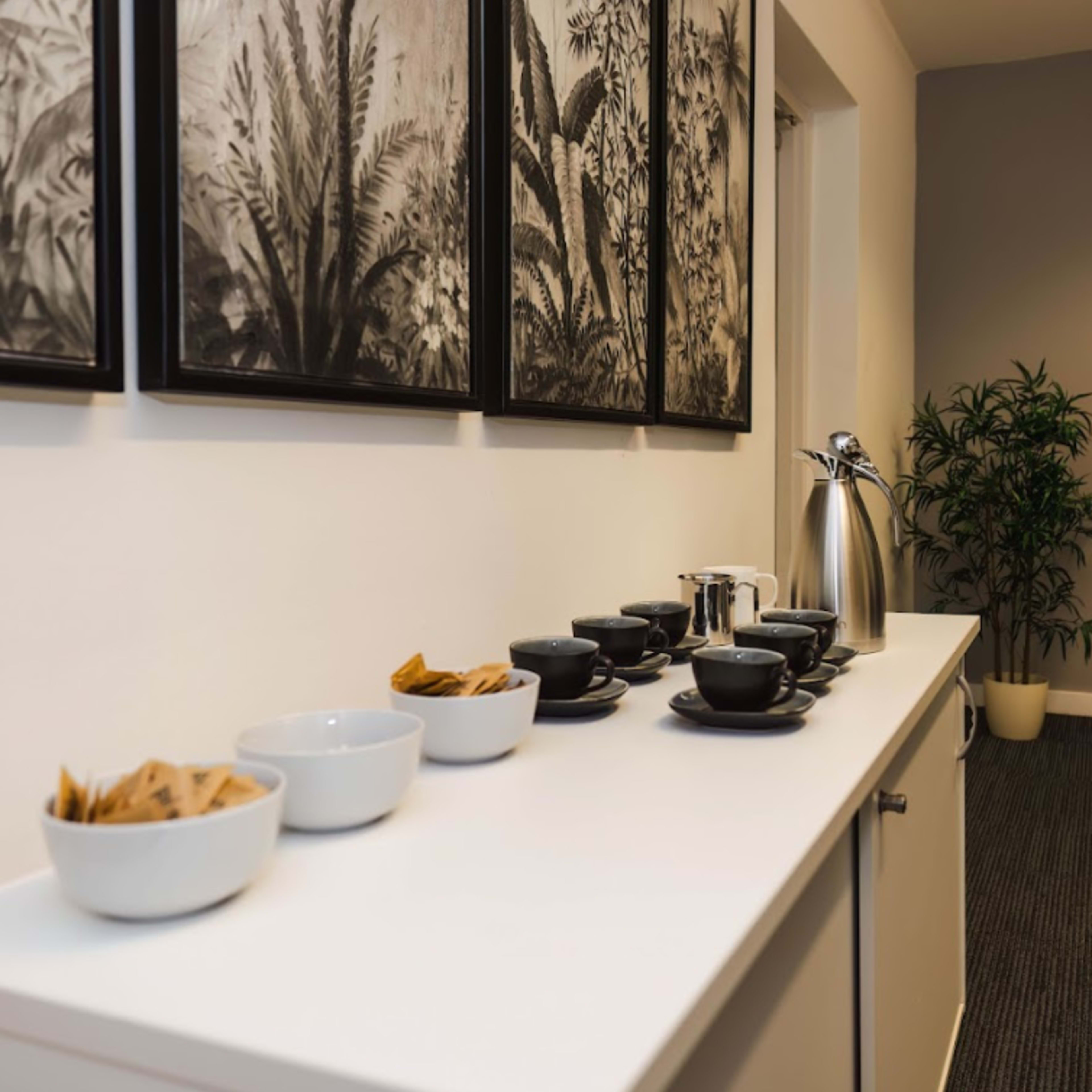 A side table features bowls of snacks, cups, and a kettle against a backdrop of framed botanical art and a potted plant.