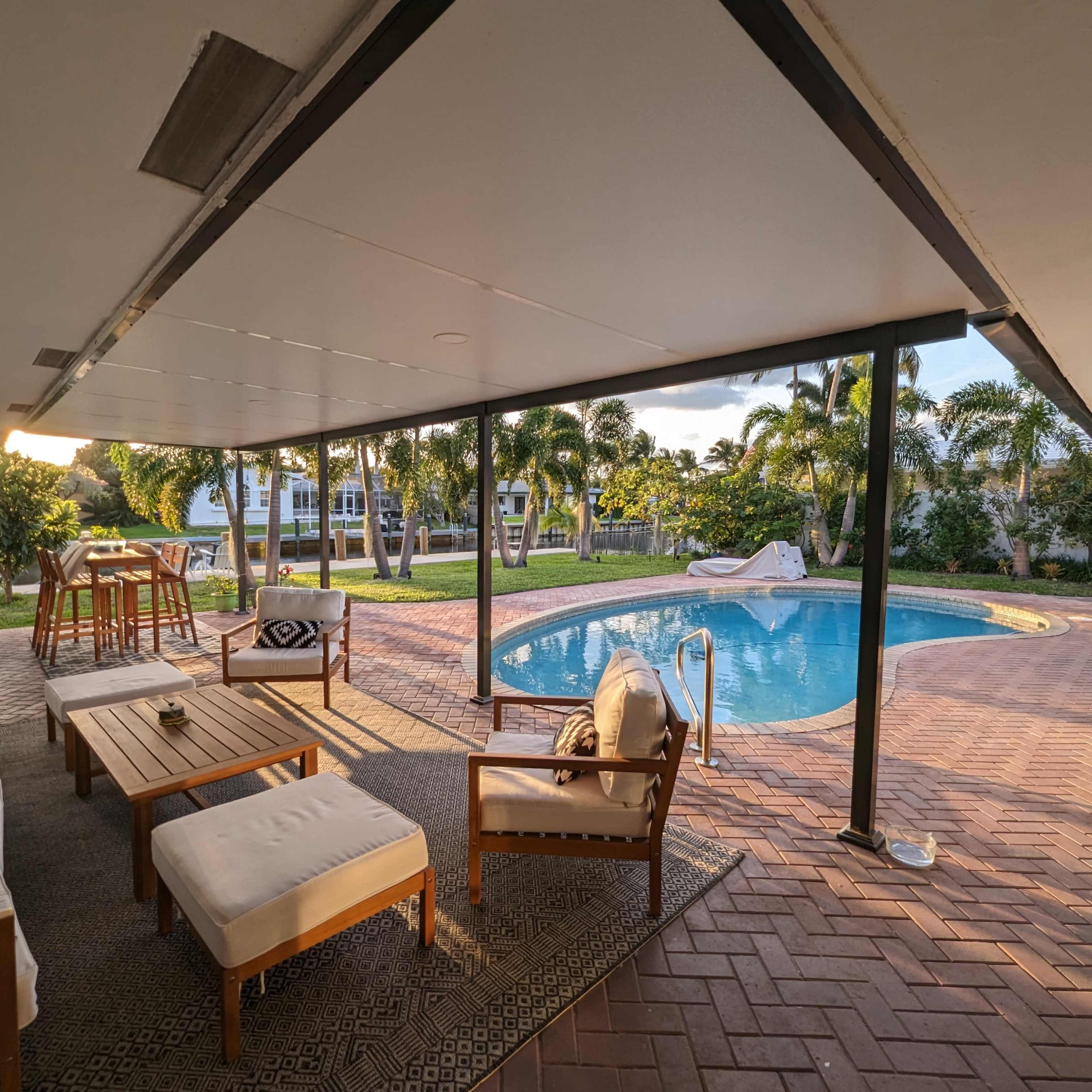 A covered patio with wooden furniture overlooks a swimming pool surrounded by palm trees and a well-maintained lawn.