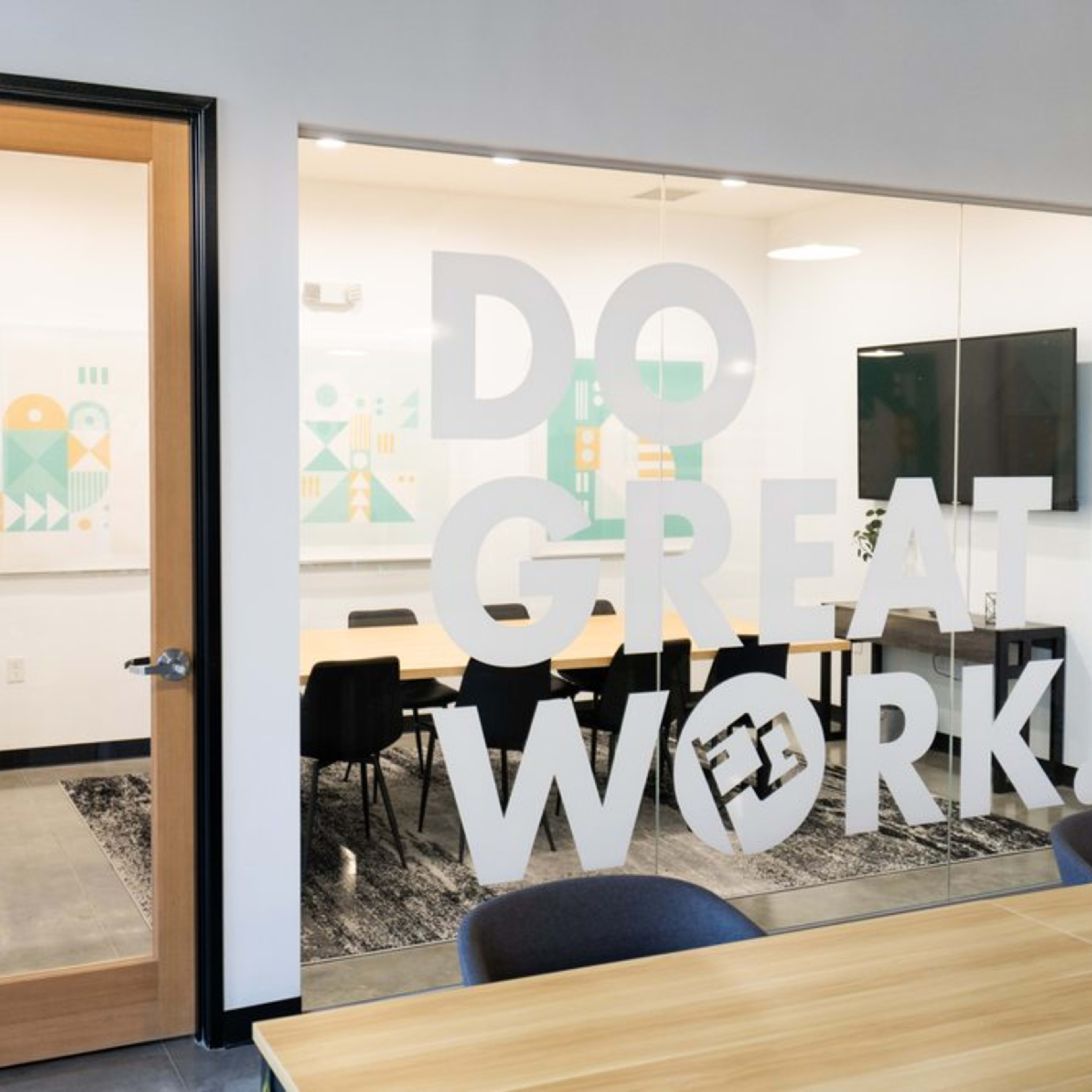 A modern conference room with a large glass wall displaying the phrase "DO GREAT WORK!" and a view of a long table surrounded by chairs.