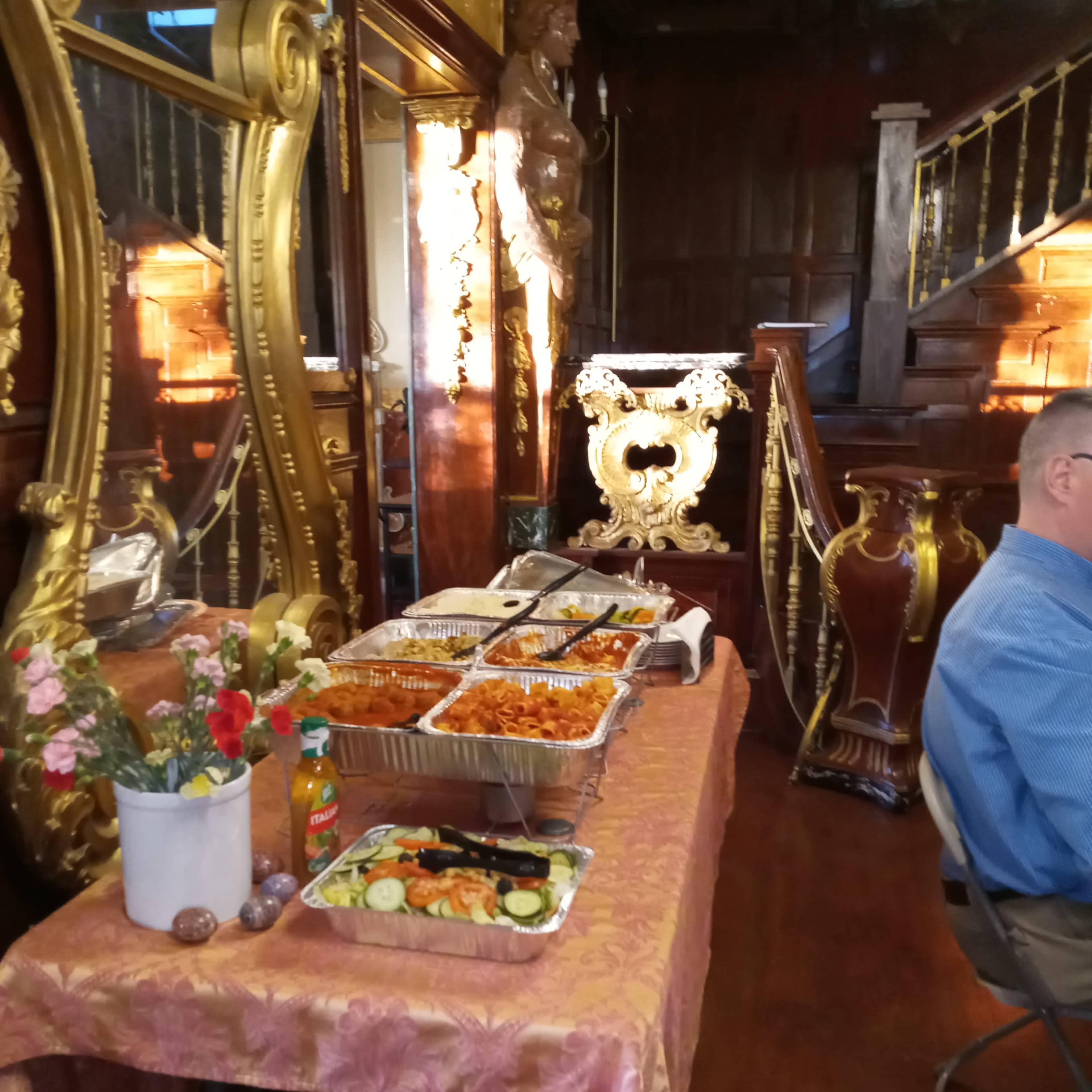 A table covered with a patterned cloth holds several trays of food, accompanied by a vase of flowers, in an ornate room featuring large mirrors and decorative wooden elements.