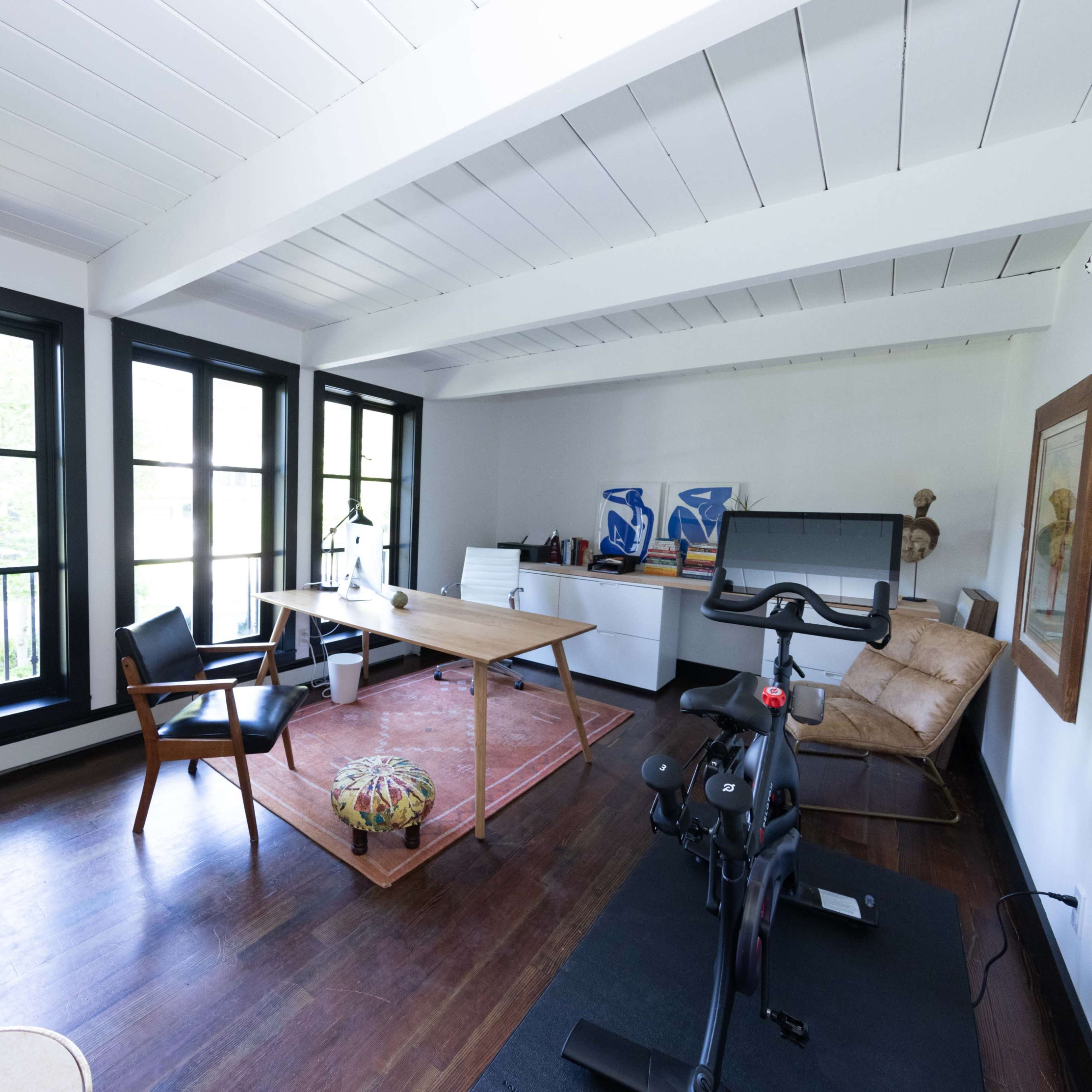 A modern home office with a wooden desk, a chair, a stationary bike, and large windows allowing natural light to enter the room.