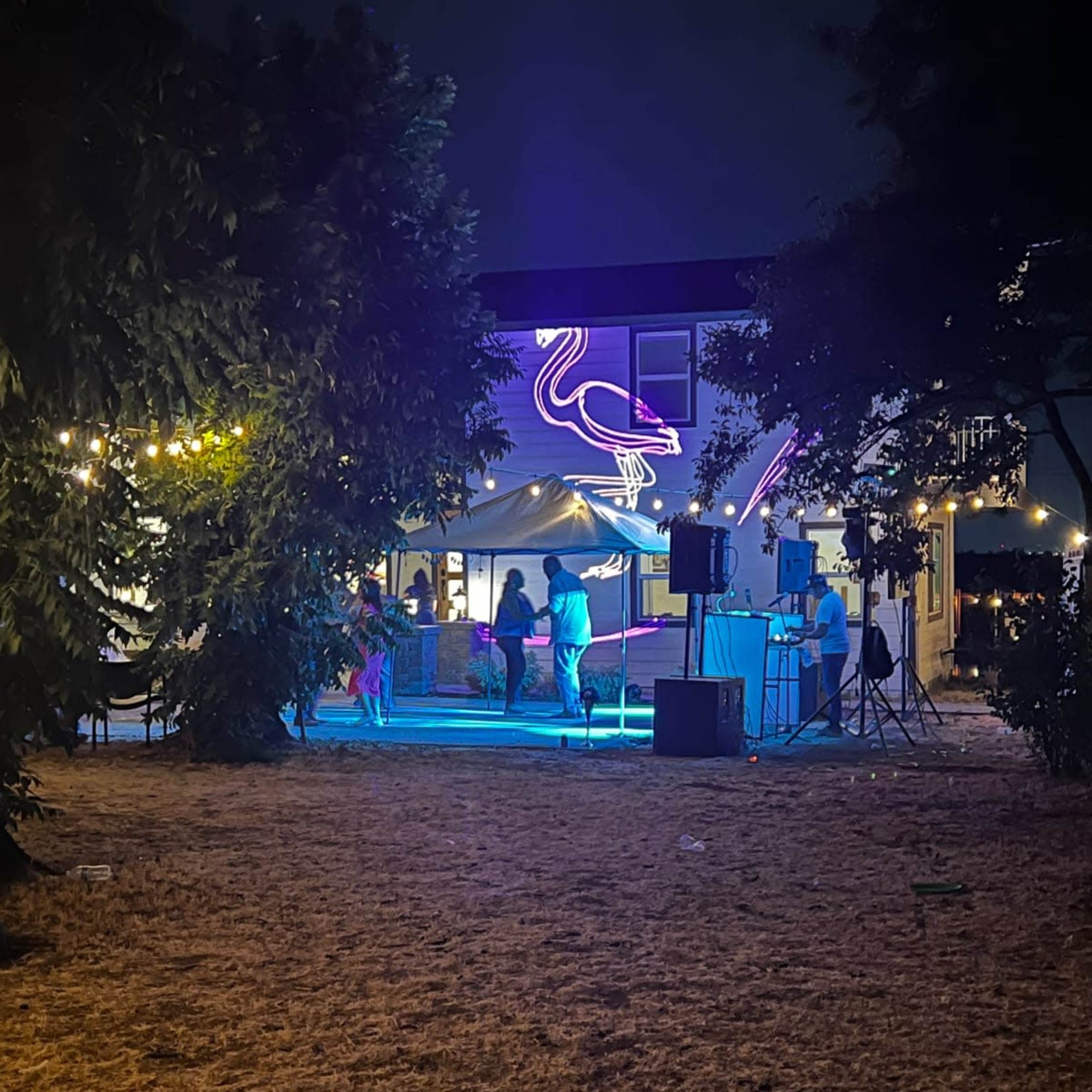 A gathering takes place outdoors at night under a neon flamingo sign, with lights illuminating a tent and a sound system set up nearby.