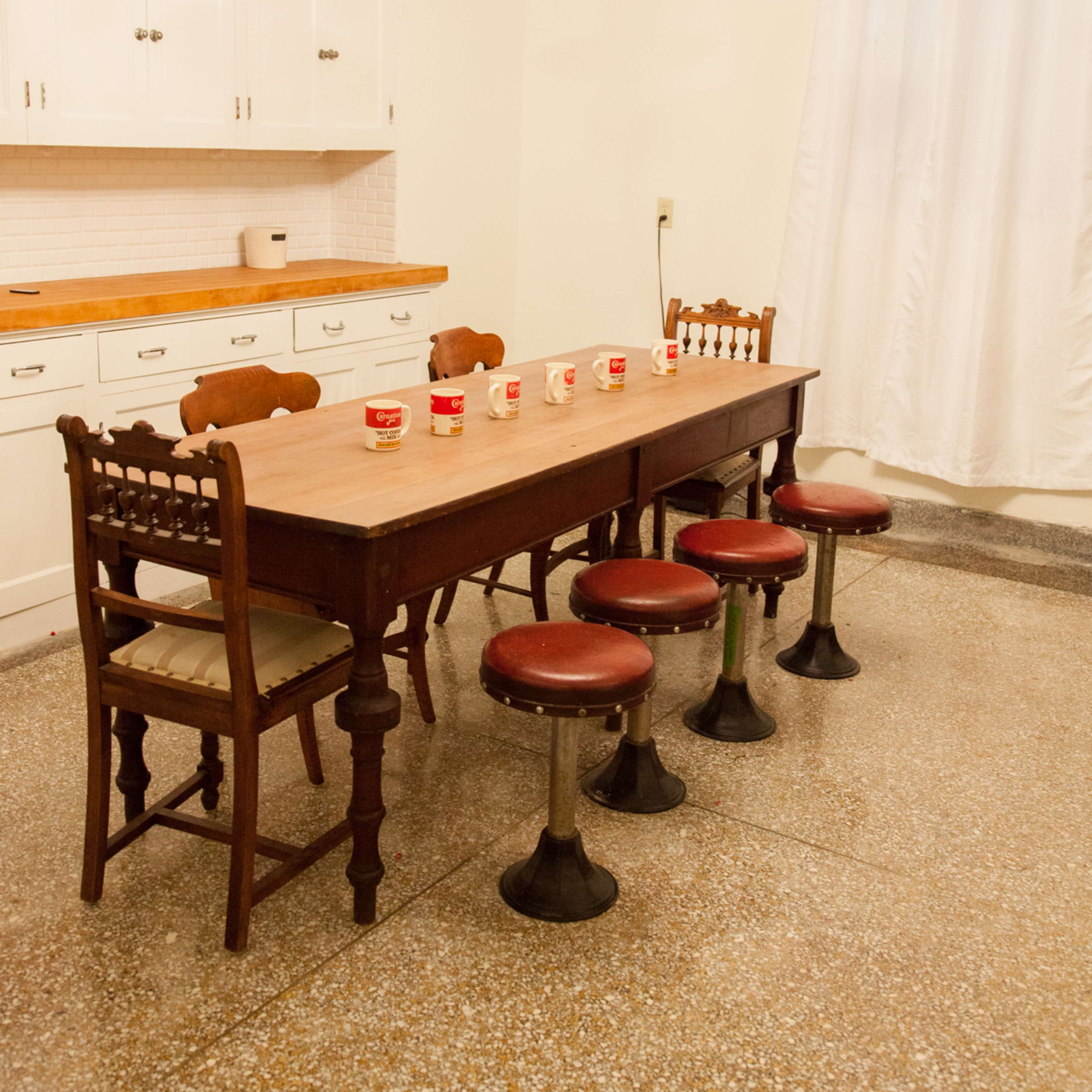A wooden dining table with six stools is set in a bright kitchen featuring white cabinets and a chandelier overhead.