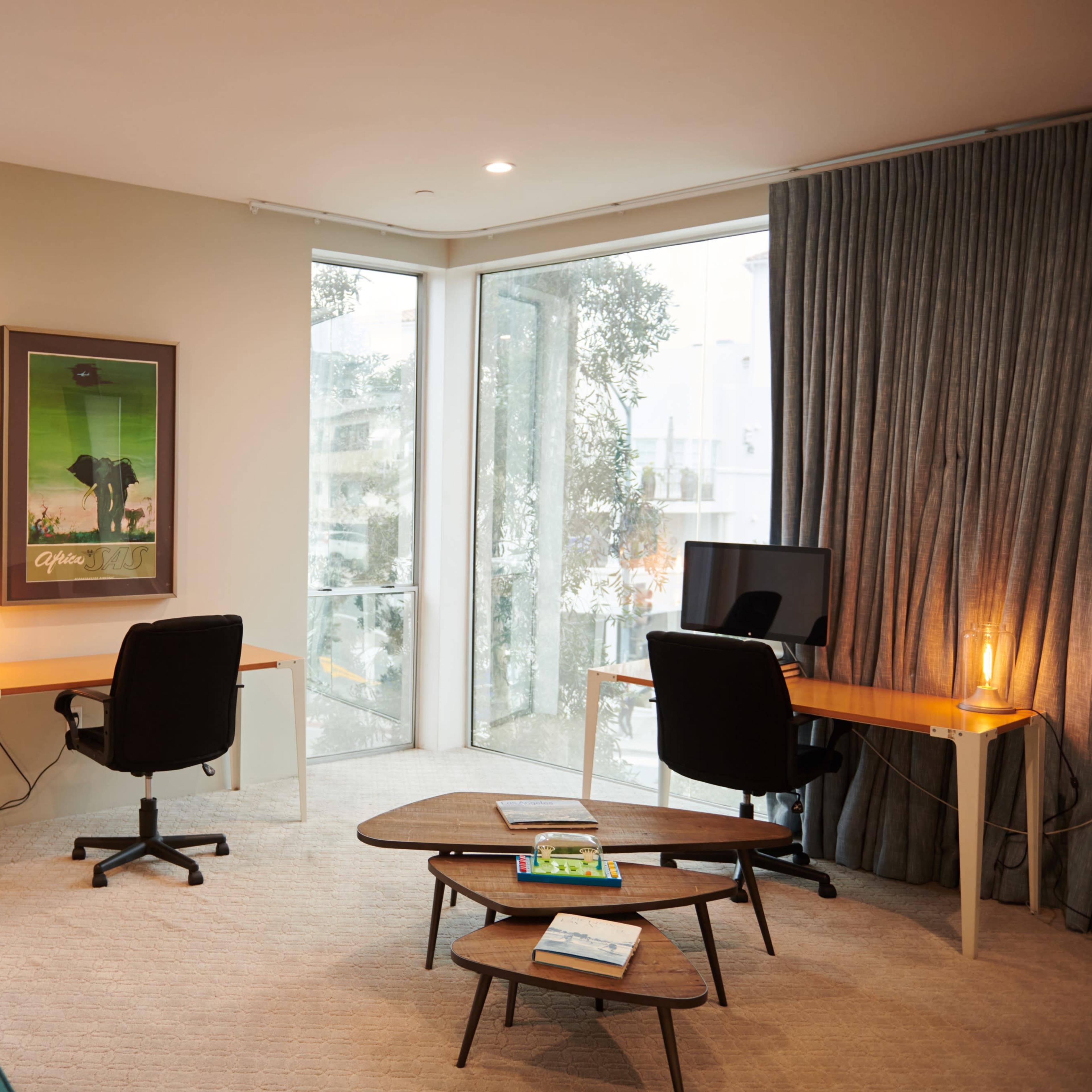 A modern living space featuring a teal sofa, two work desks with chairs, large windows, and a coffee table.