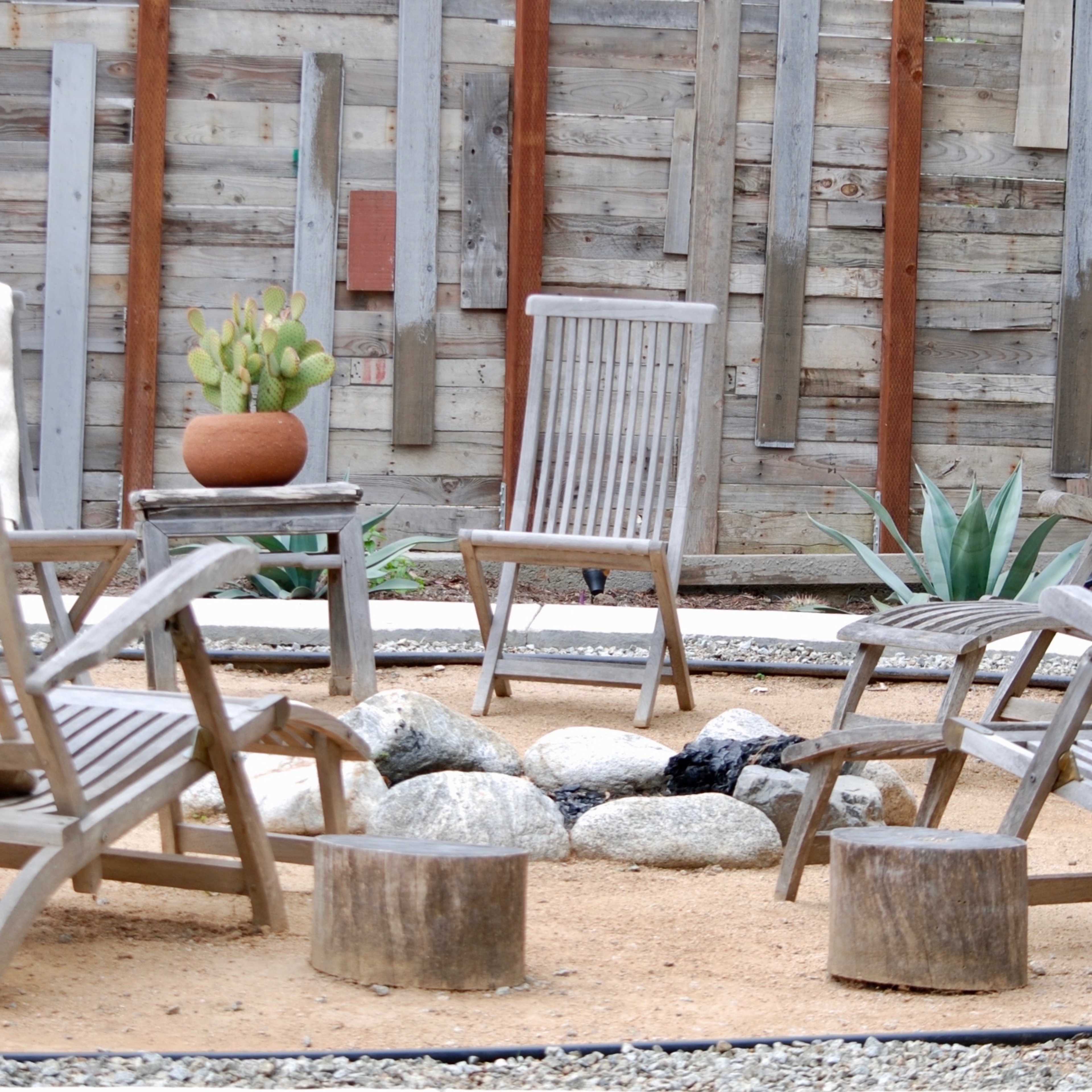 A circle of wooden chairs arranged around a fire pit in a landscaped yard with gravel and plants.