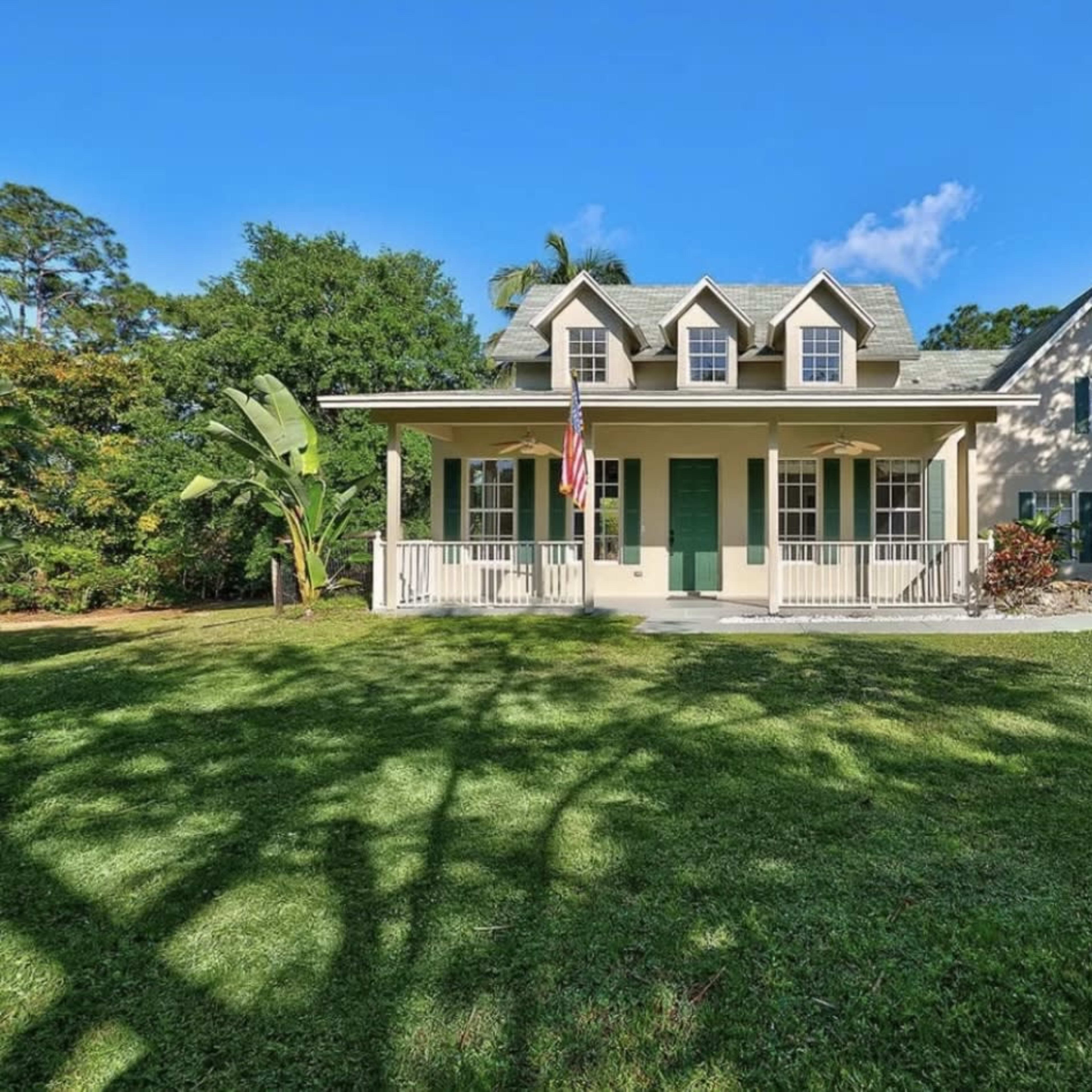 A two-story house with green shutters and an American flag stands amidst a grassy lawn surrounded by trees.