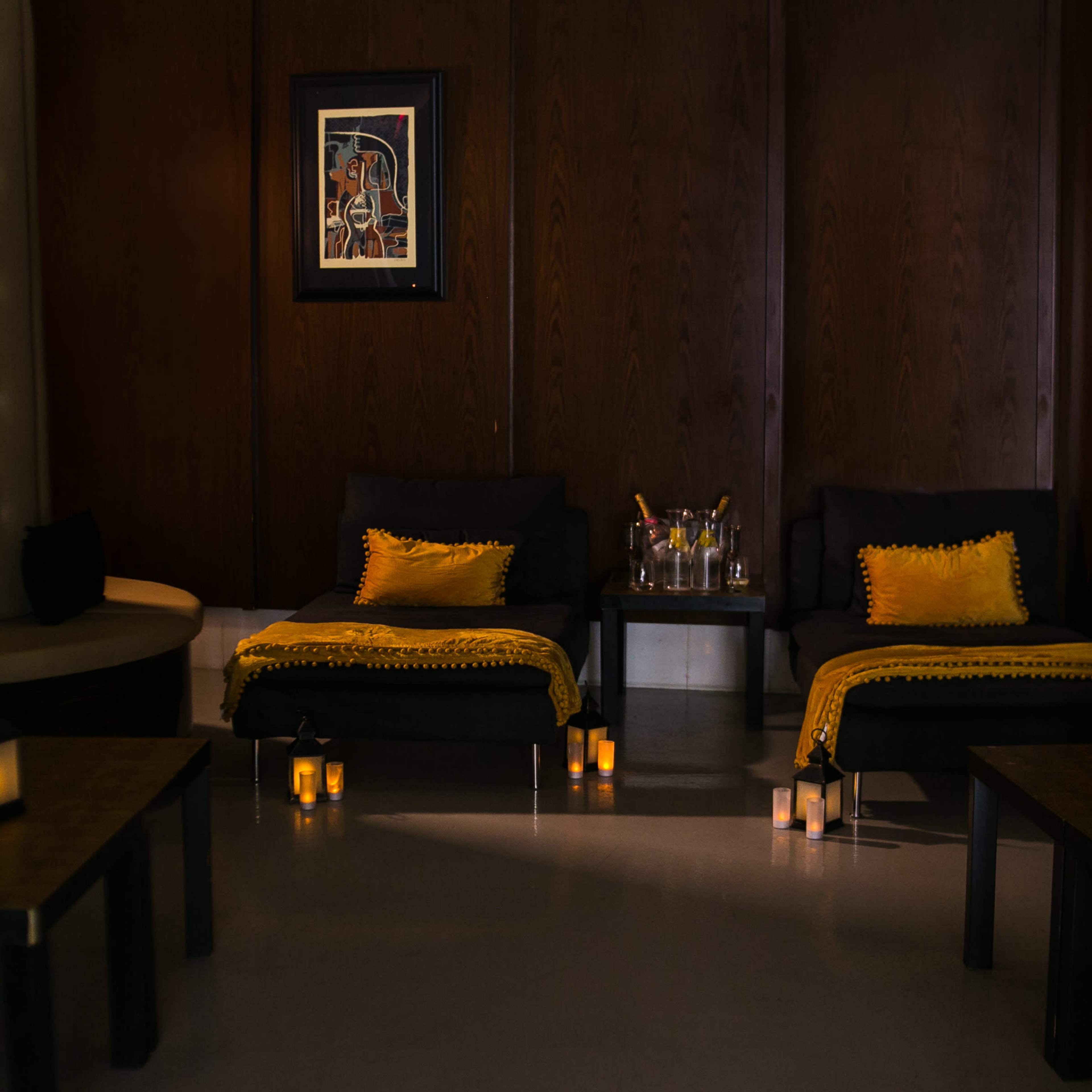 A dimly lit relaxation area featuring two lounge chairs with yellow throws, a small table with bottles and glasses, and several lit candles.