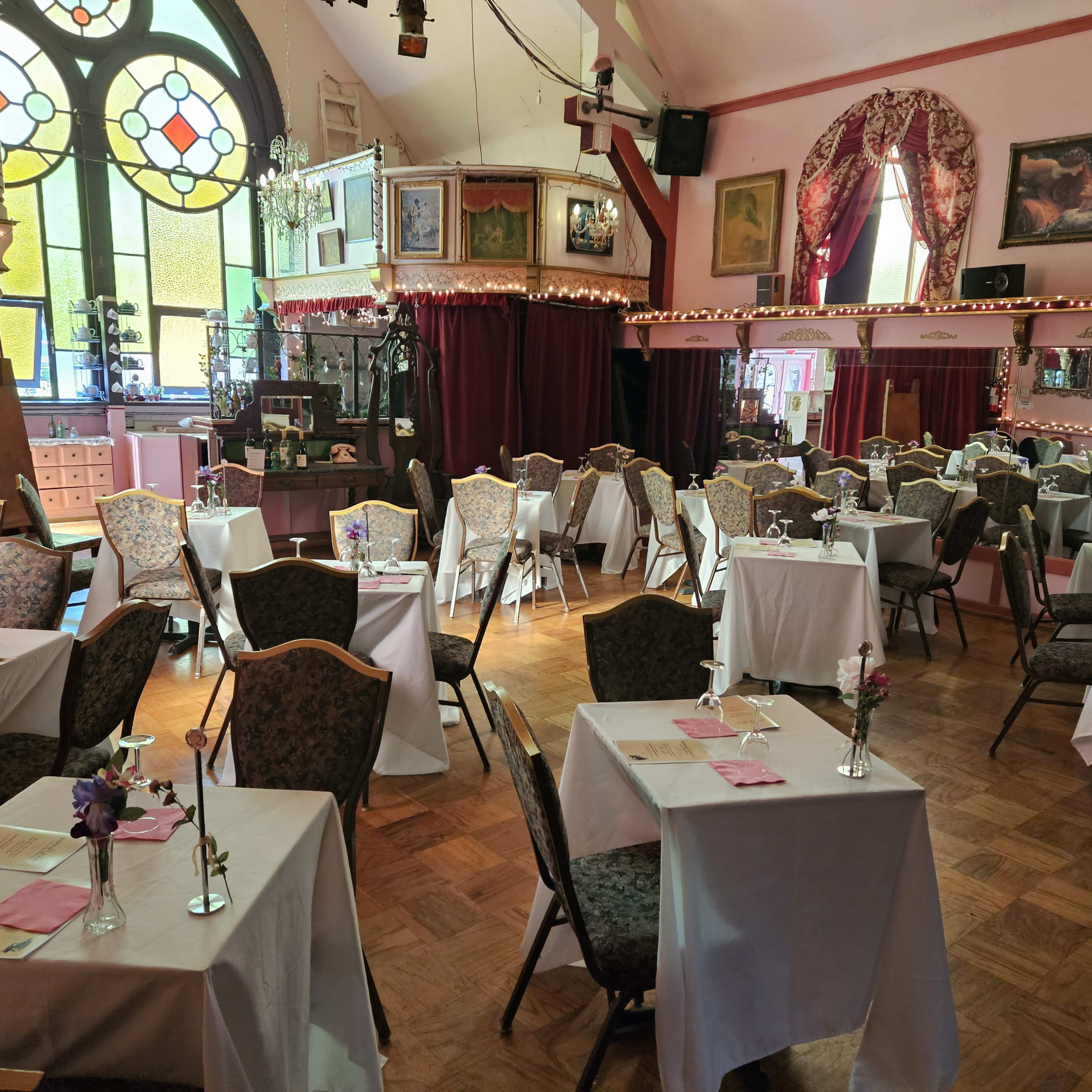 A dining area with tables covered in white tablecloths, decorated with small vases of flowers, and surrounded by framed artwork and stained glass windows.