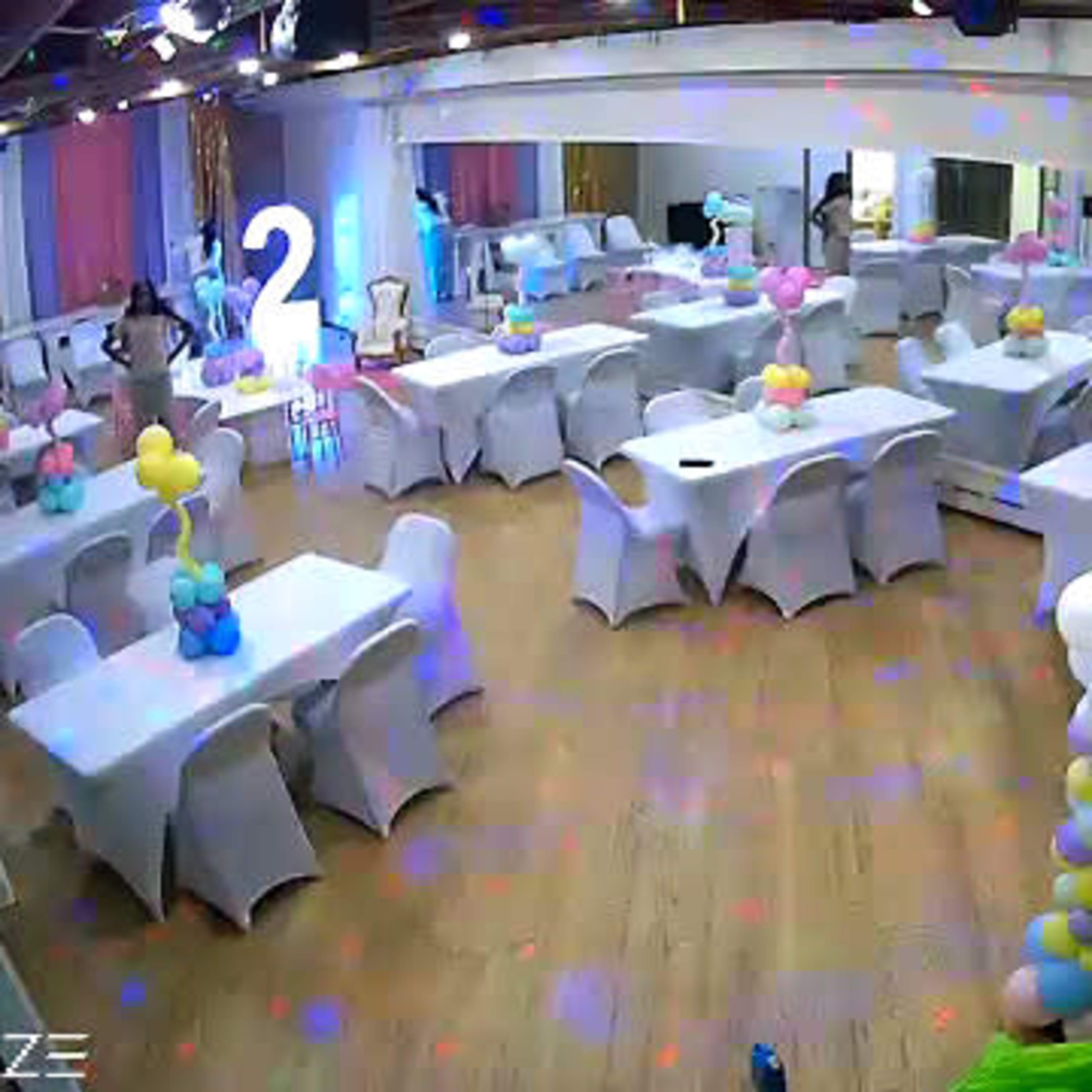 A decorated event room with white tables and chairs, colorful balloon arrangements, and a large illuminated number "2" in the background.