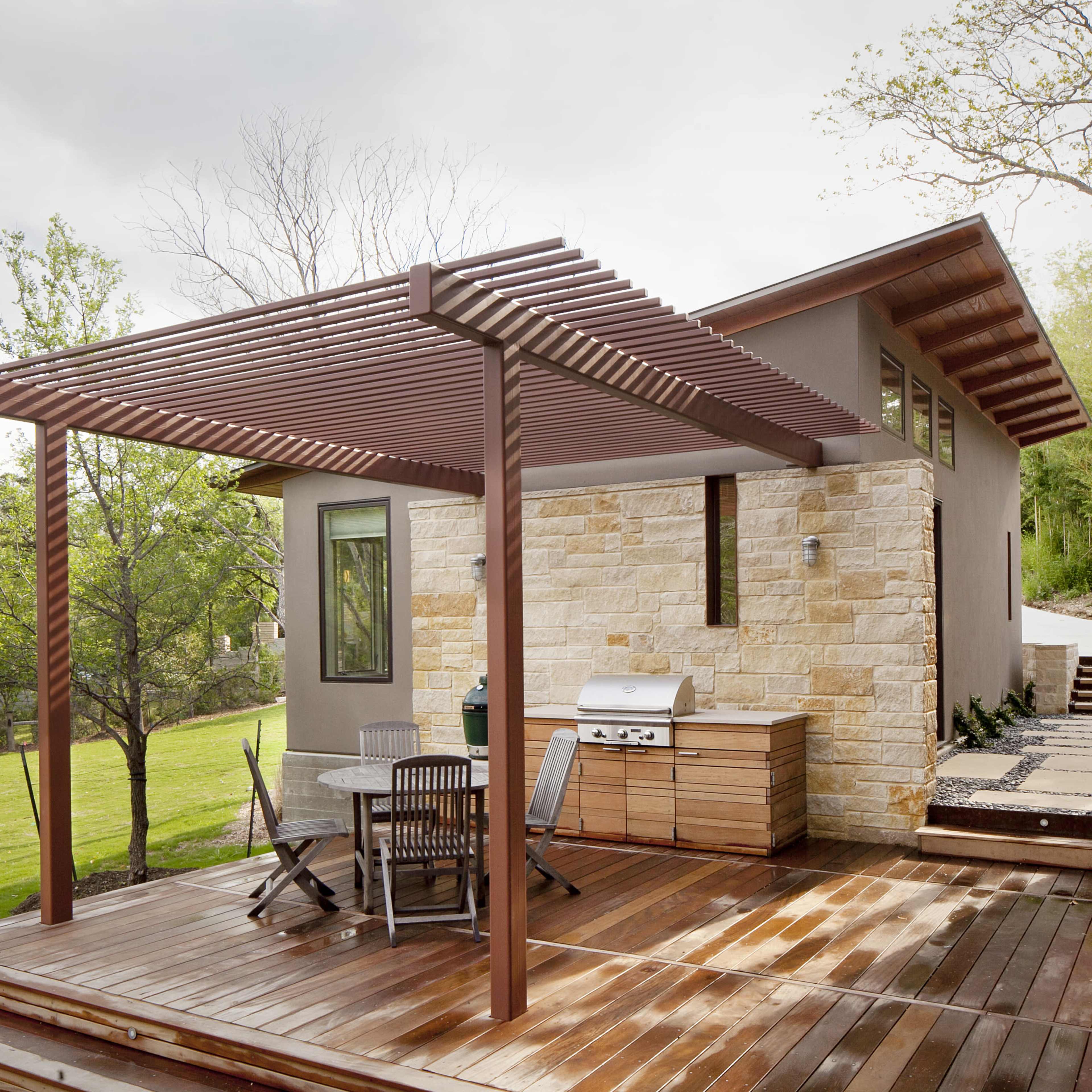 A wooden deck features a dining set and a barbecue grill under a slatted pergola adjacent to a modern house.