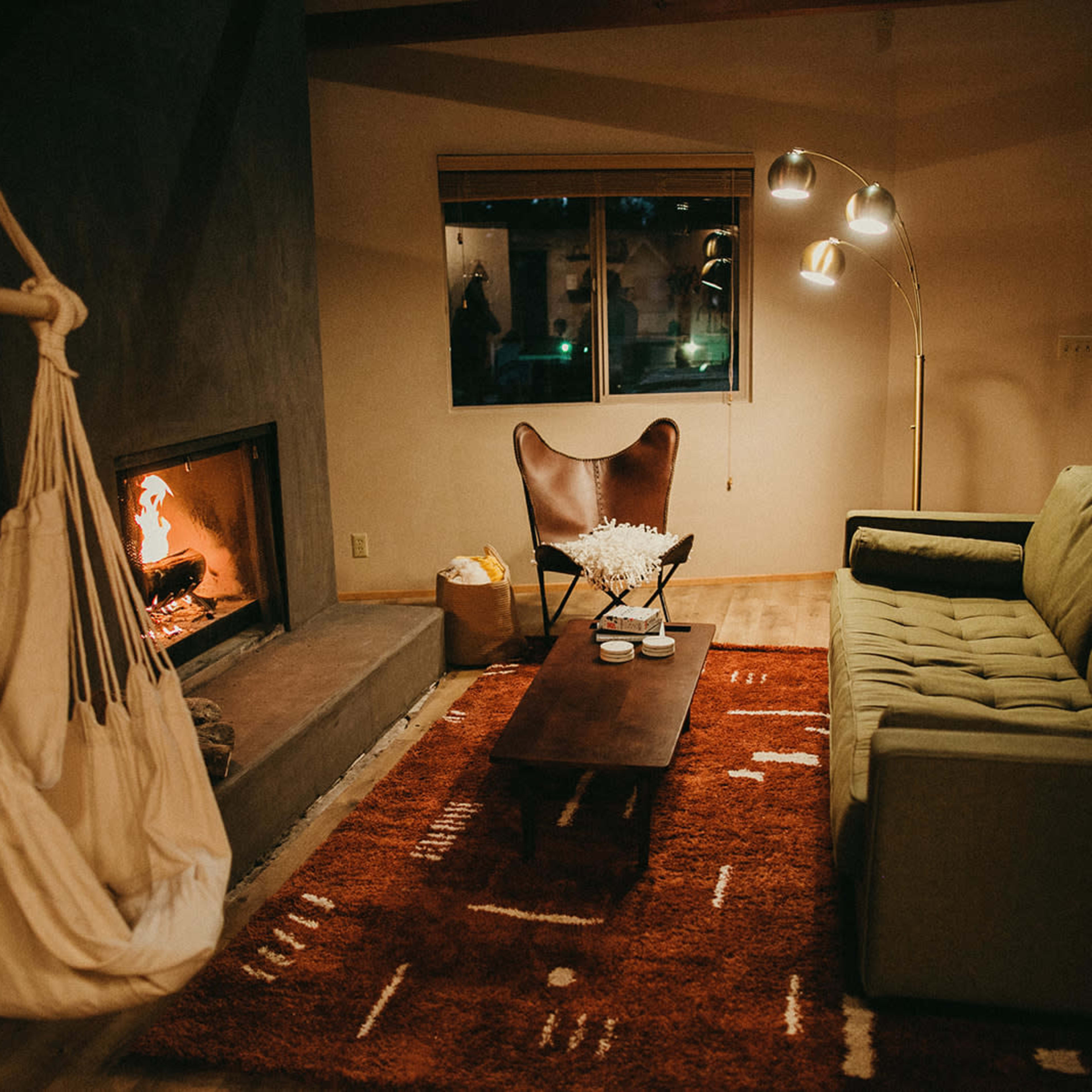 The living room features a green sofa, a wooden coffee table, a hanging chair, and a fireplace, all set against a backdrop of warm lighting and a rustic rug.