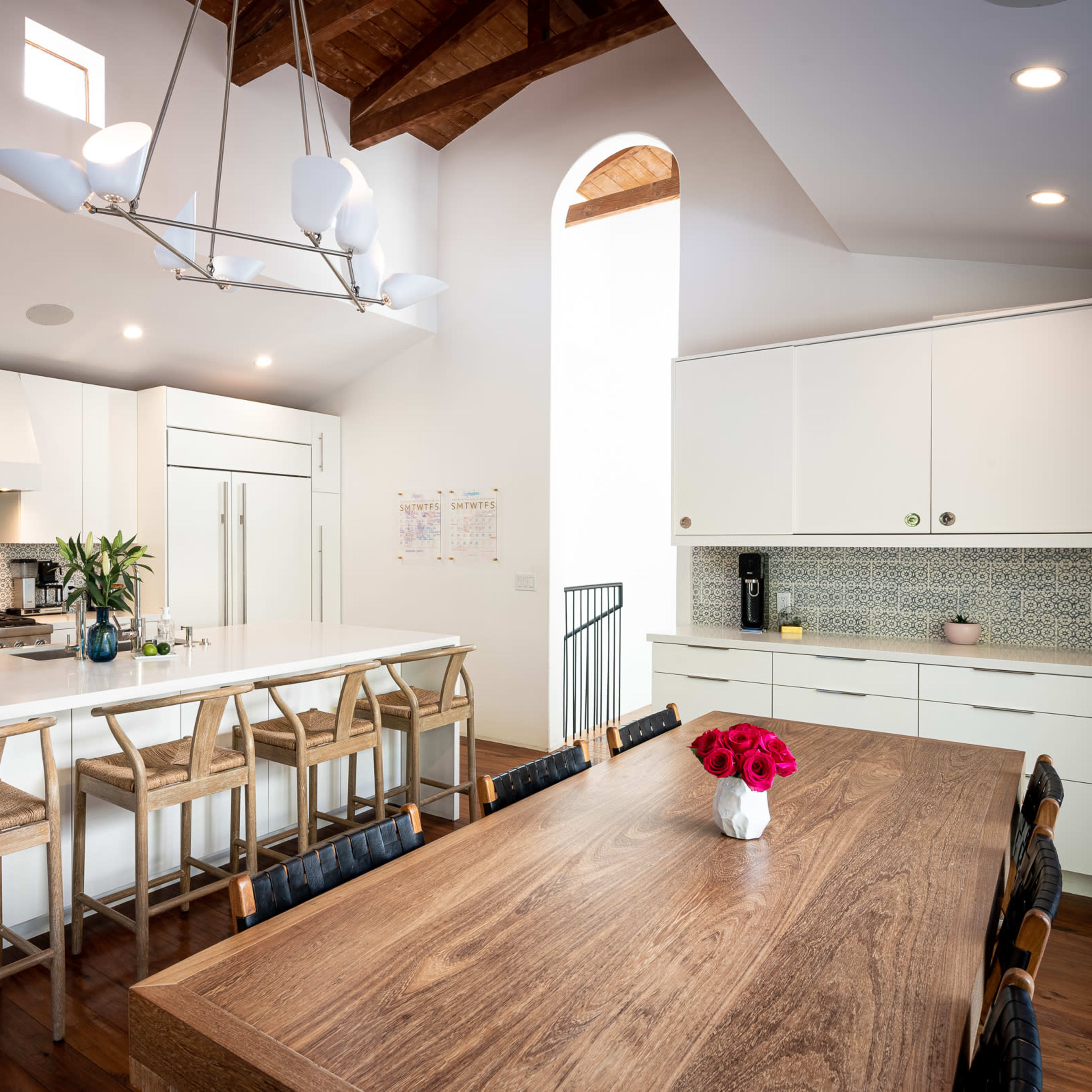 A spacious kitchen features a large island with bar stools, white cabinetry, and a dining table adorned with a vase of flowers.