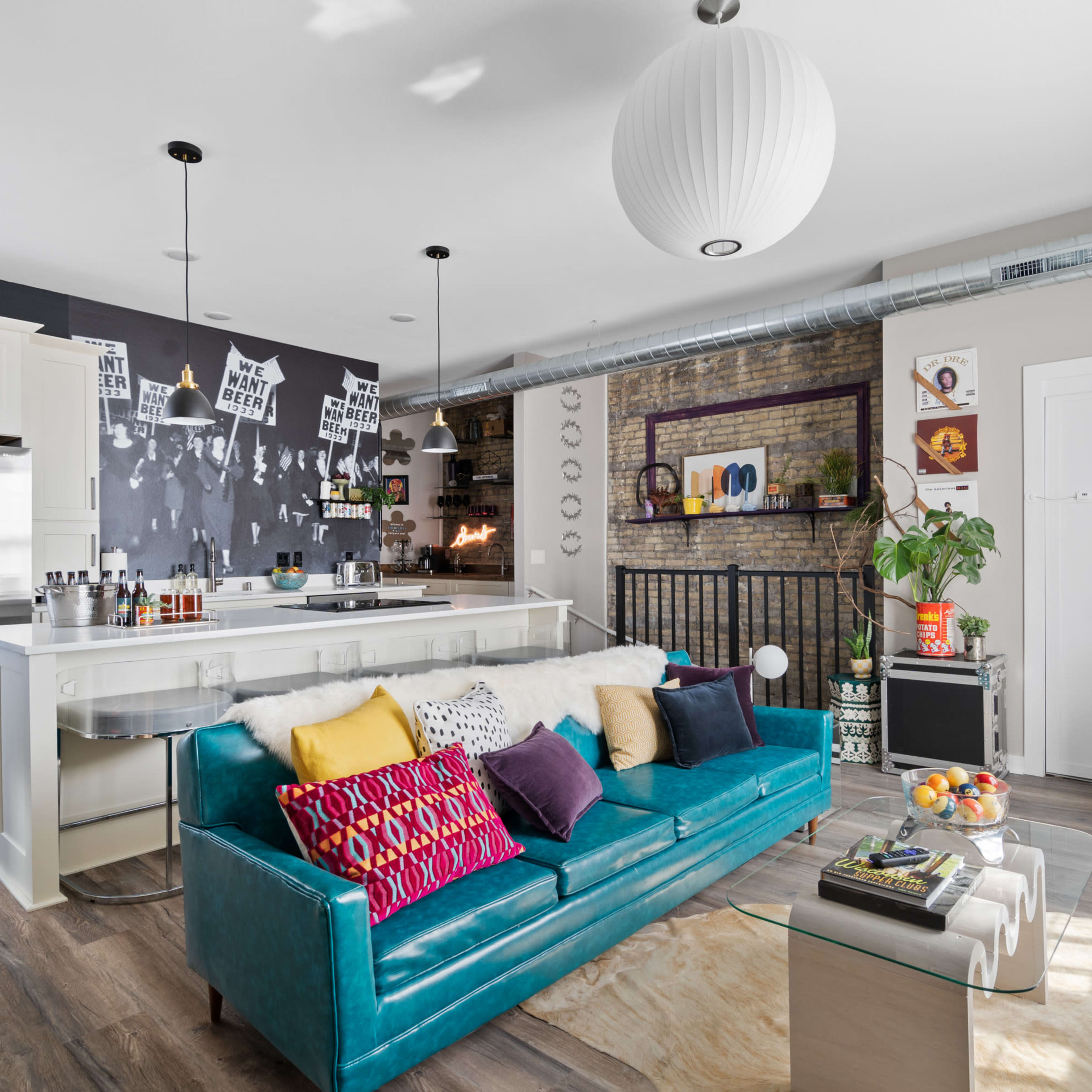 A modern living room features a teal couch, a white kitchen island, an exposed brick wall with decor, and various decorative elements throughout the space.