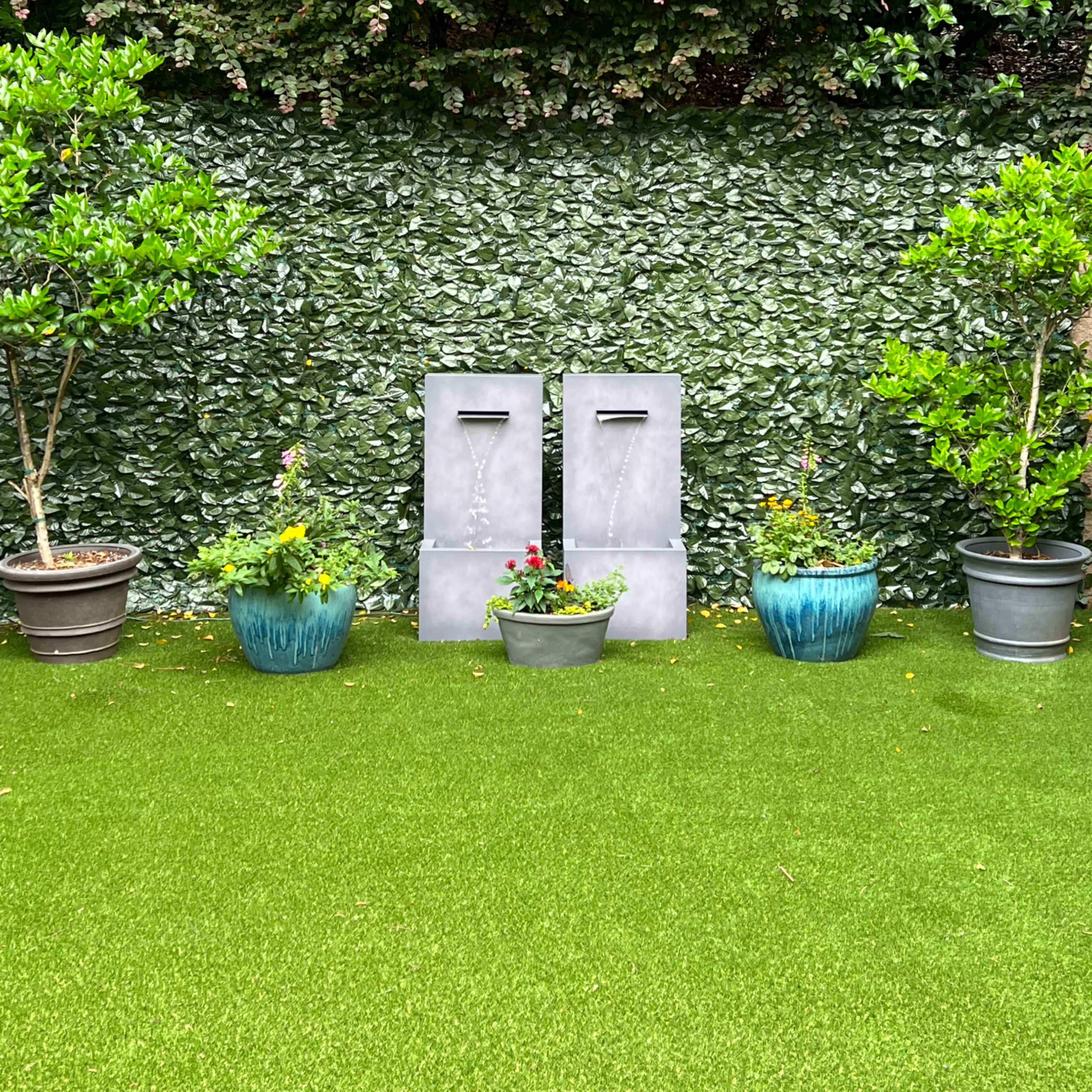 A green artificial grass area with two gray rectangular sculptures and potted plants against a leafy backdrop.