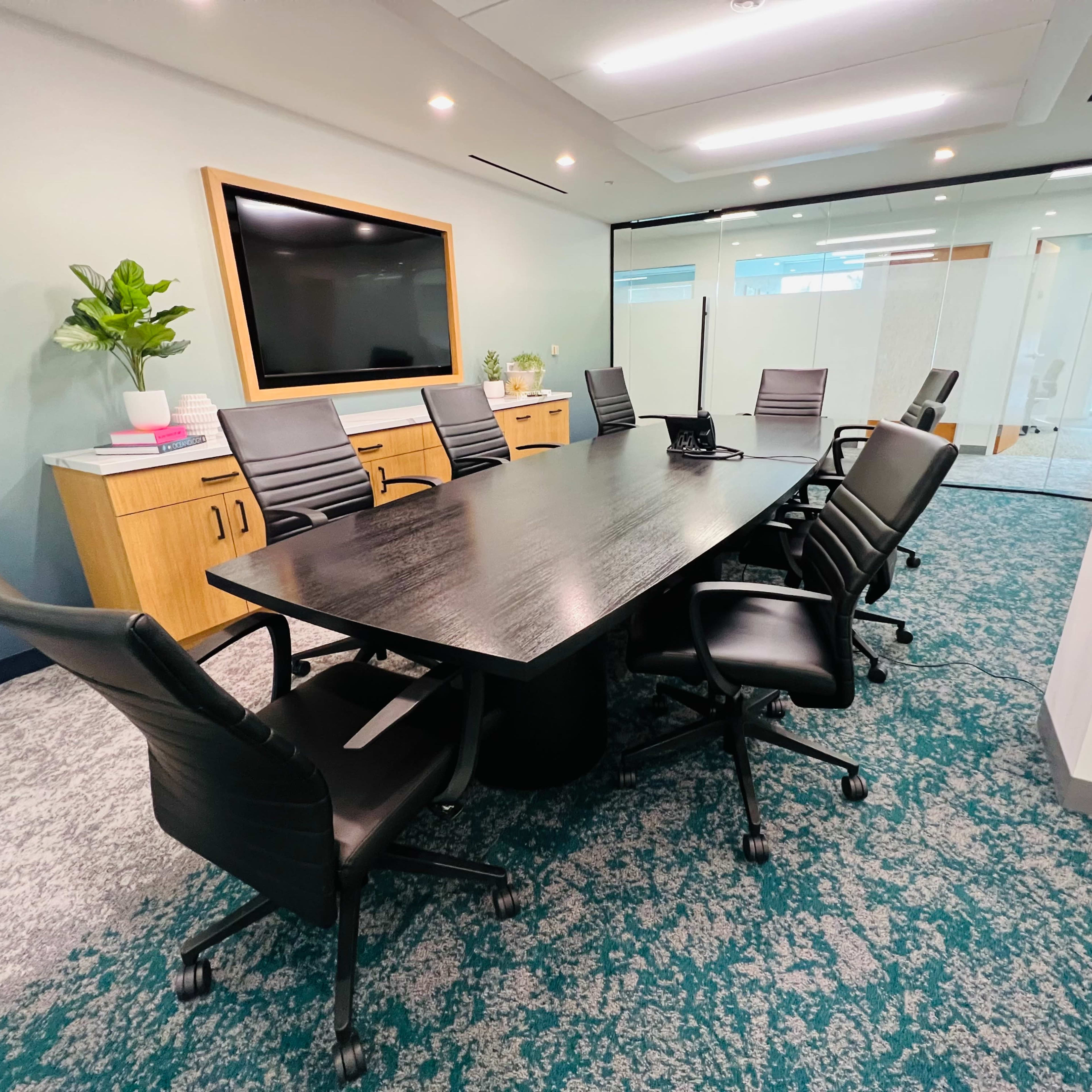A modern conference room features a long black table surrounded by ergonomic chairs, with a large television screen mounted on the wall and a view of glass-walled offices.