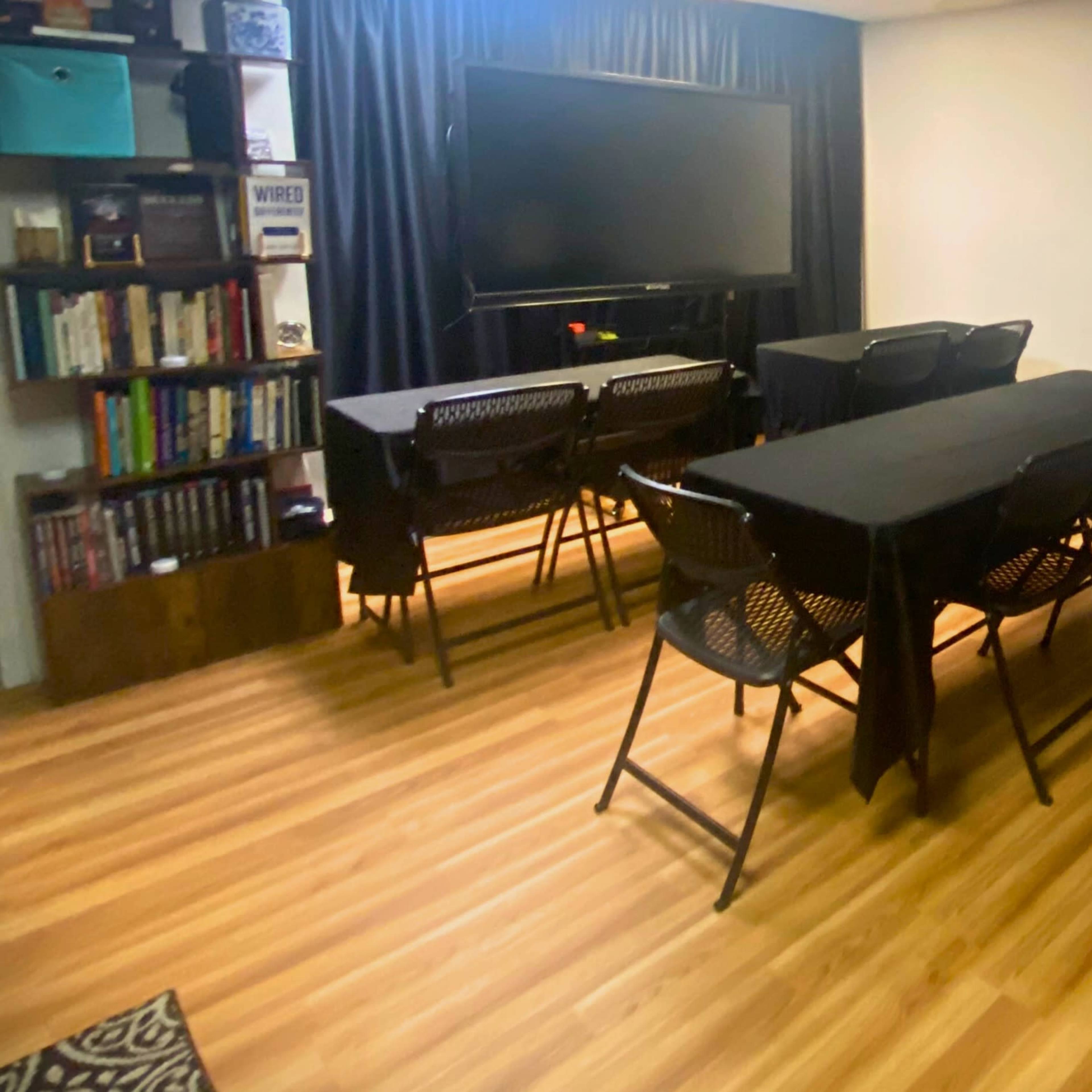 The room features several black folding tables arranged with chairs, a large television on the wall, and a bookshelf in the background.