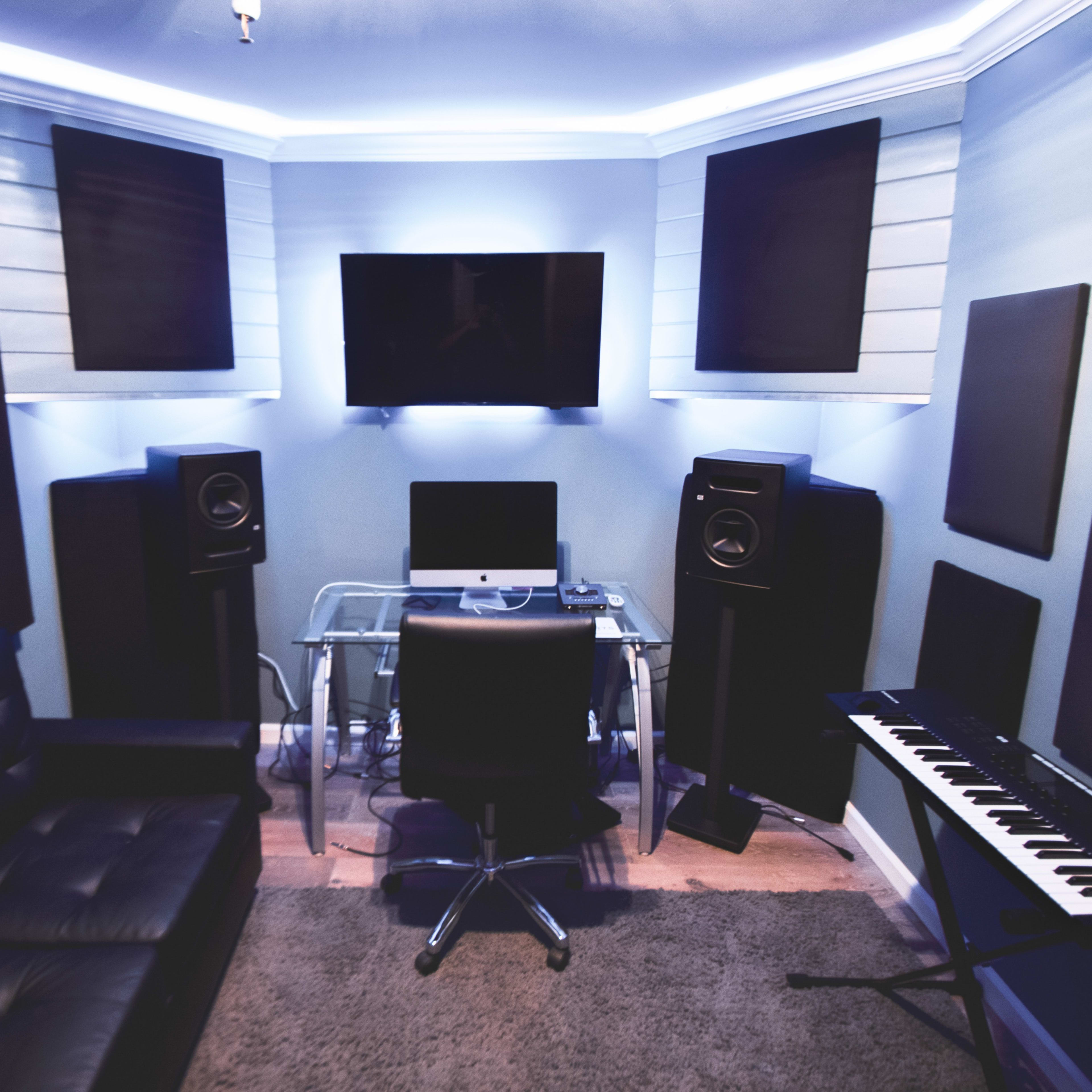 A music production studio featuring a glass desk, an office chair, audio monitors, and a keyboard, with soundproofing panels on the walls and a television mounted above.