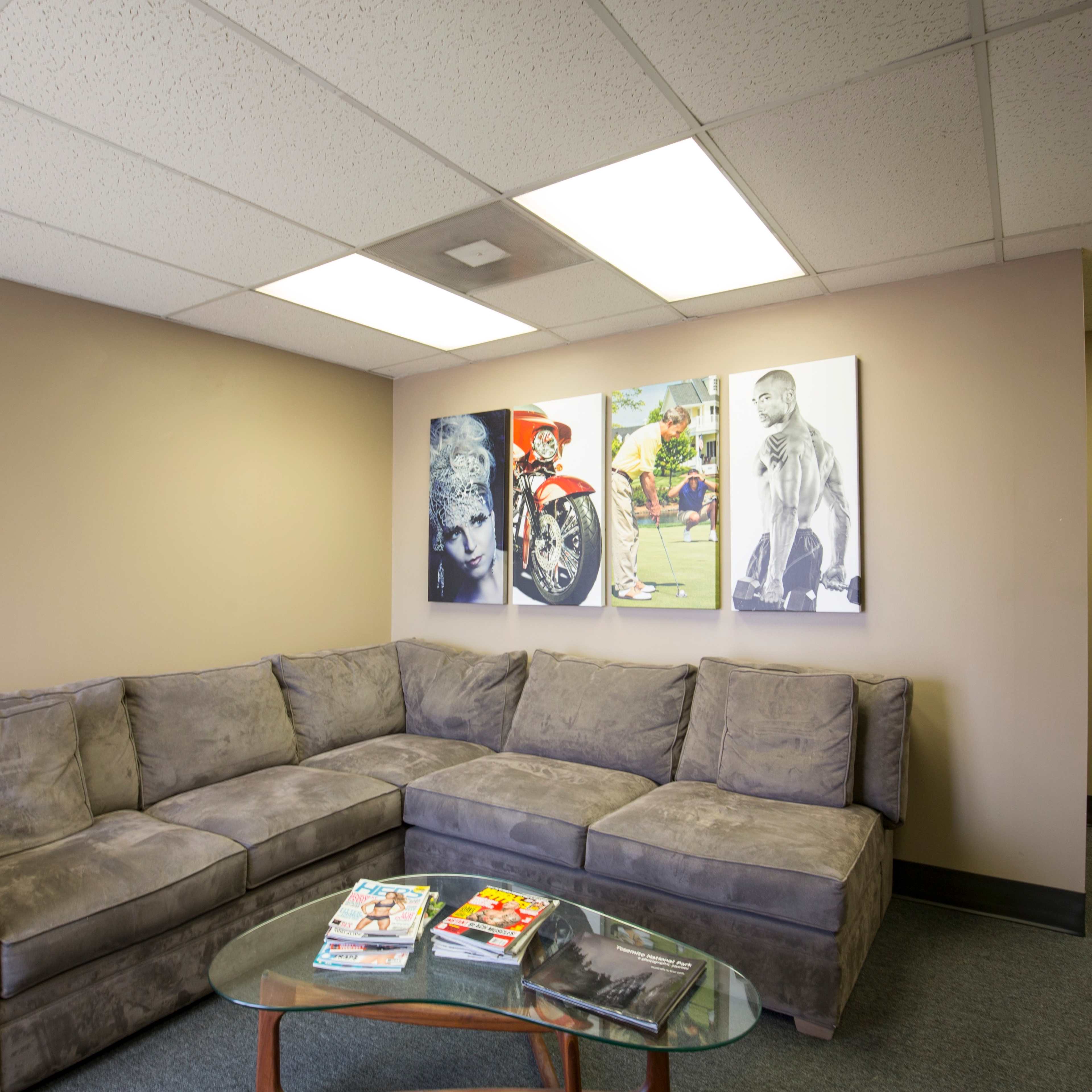 A cozy lounge area featuring a gray sectional sofa, a glass coffee table with magazines, and three large framed pictures on the wall.