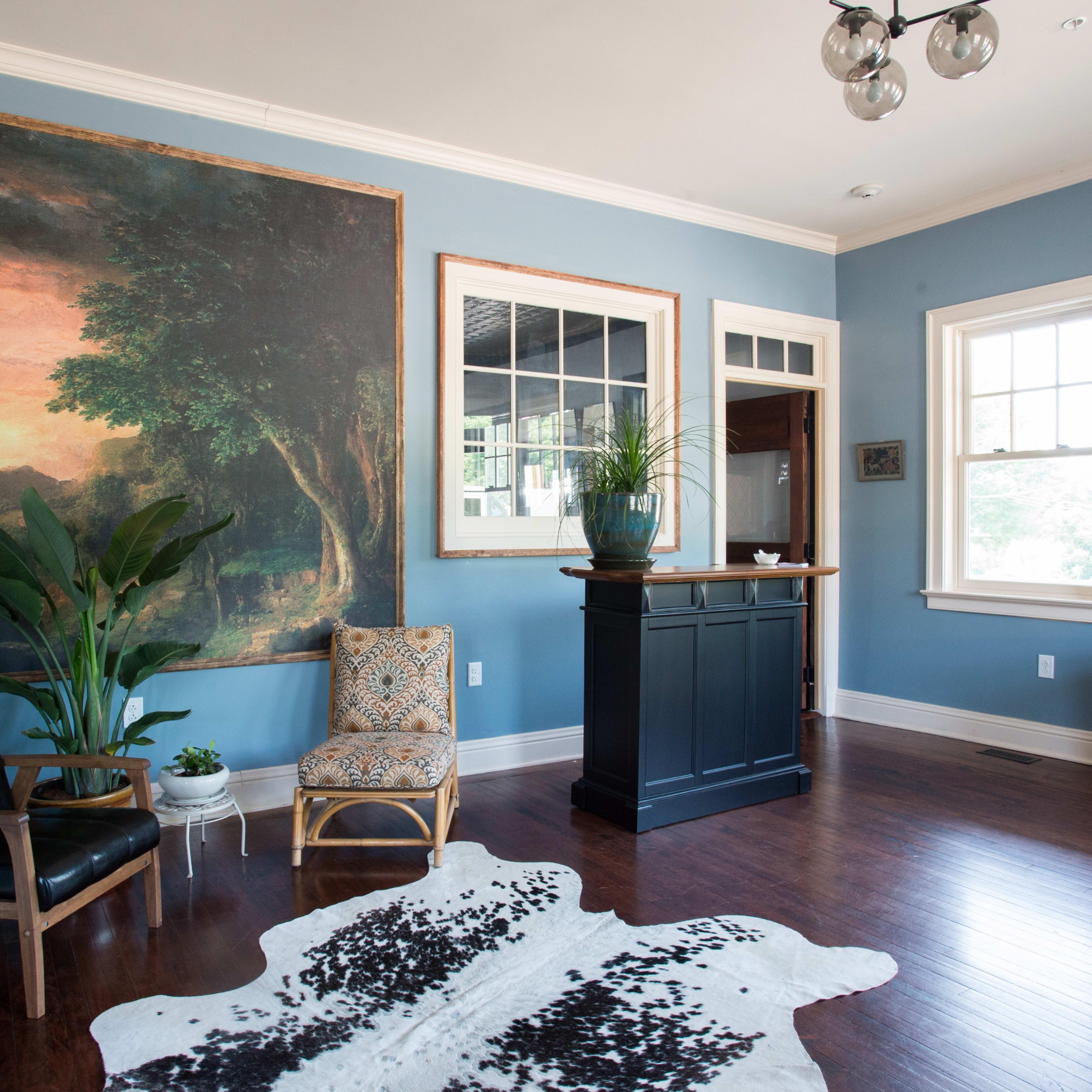 The room features blue walls, a large landscape painting, a patterned armchair, a sleek black chair, and a small bar area with plants and natural light.