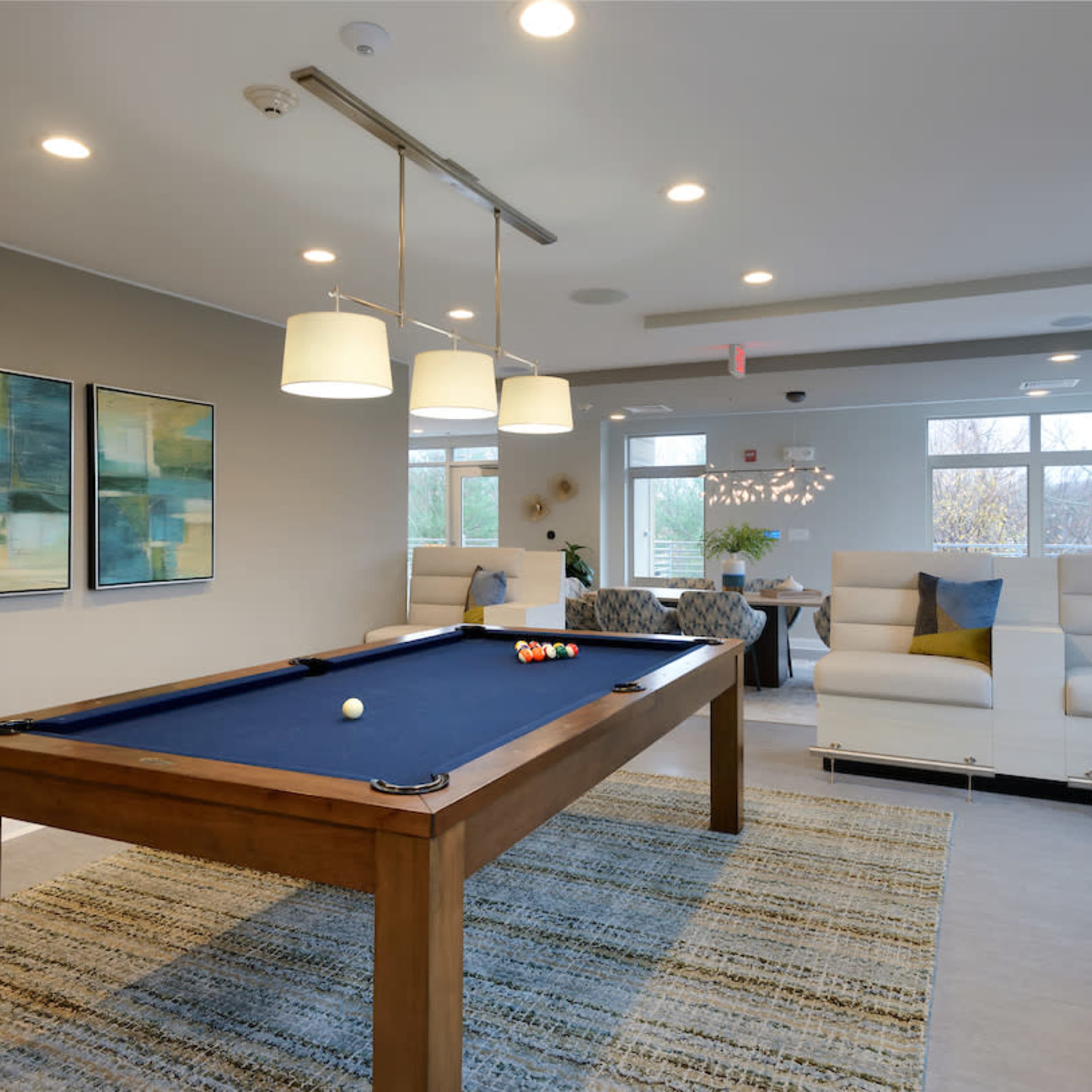 A modern game room featuring a billiards table with a blue felt surface, surrounded by a seating area and large windows.