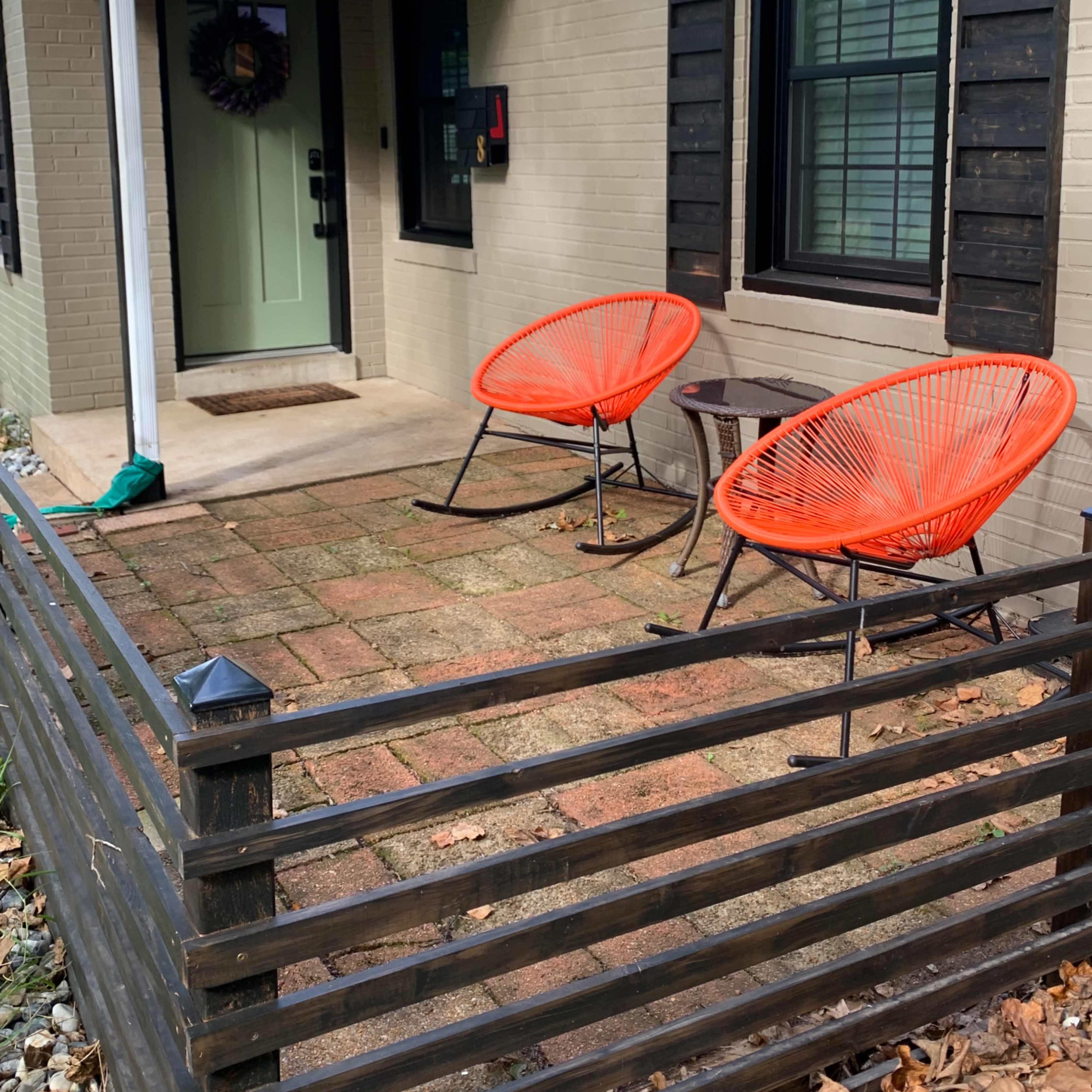 A small front porch with two orange chairs and a side table, bordered by a wooden fence.