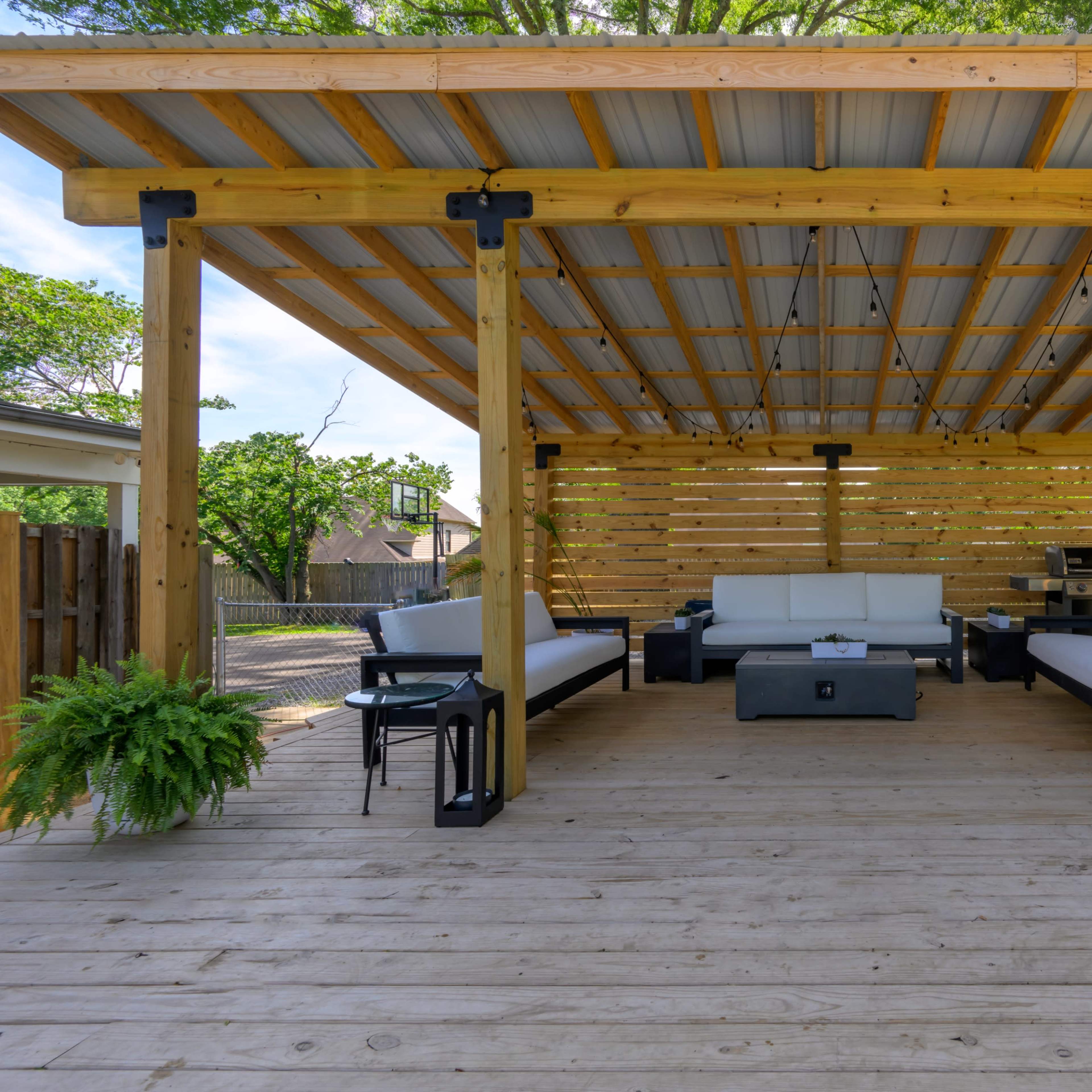 A wooden deck features a covered seating area with a sofa set, coffee table, and planters.